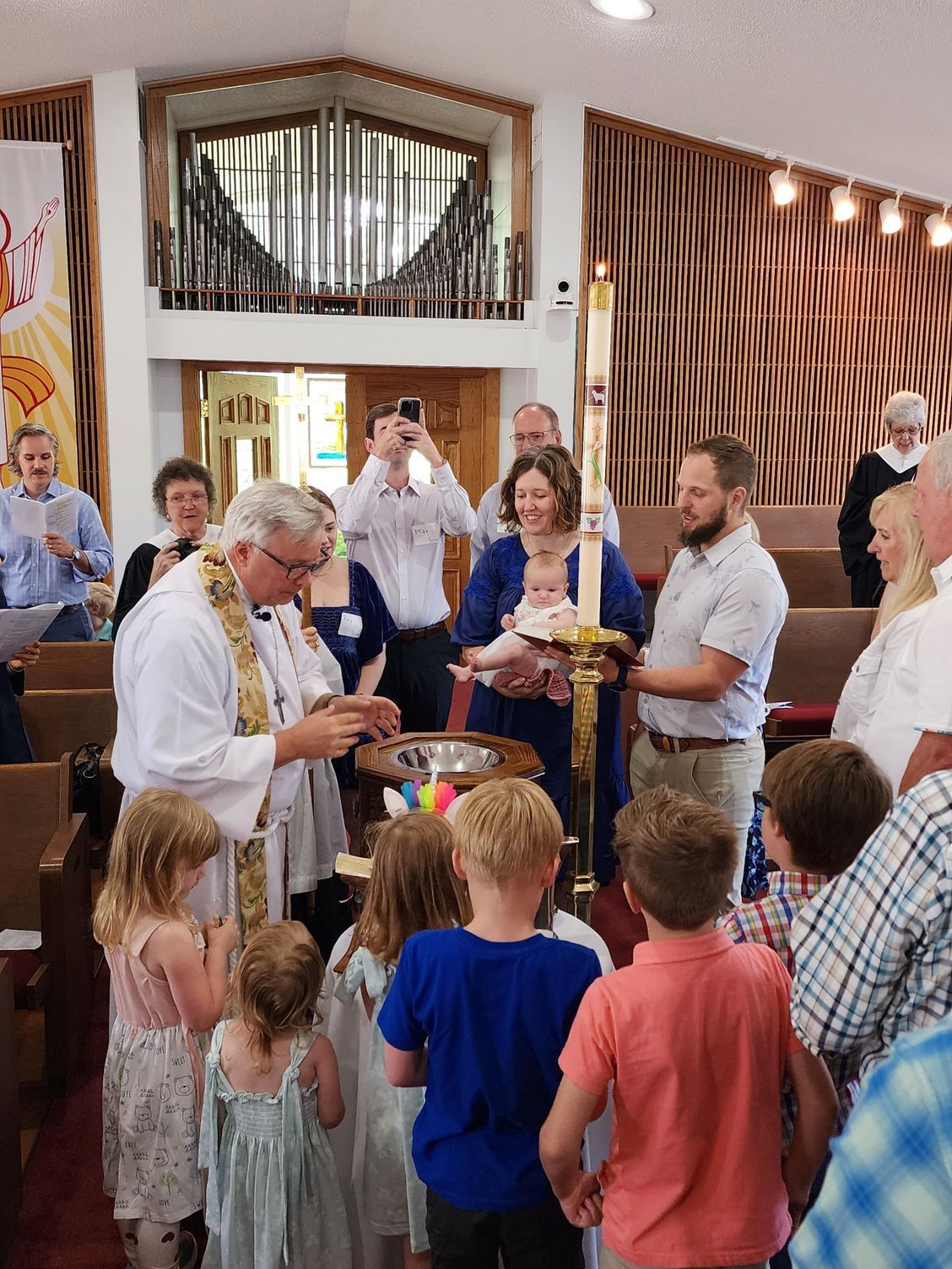 Baptism ceremony in a church. Priest pours water over a baby held by parents. Children watch.