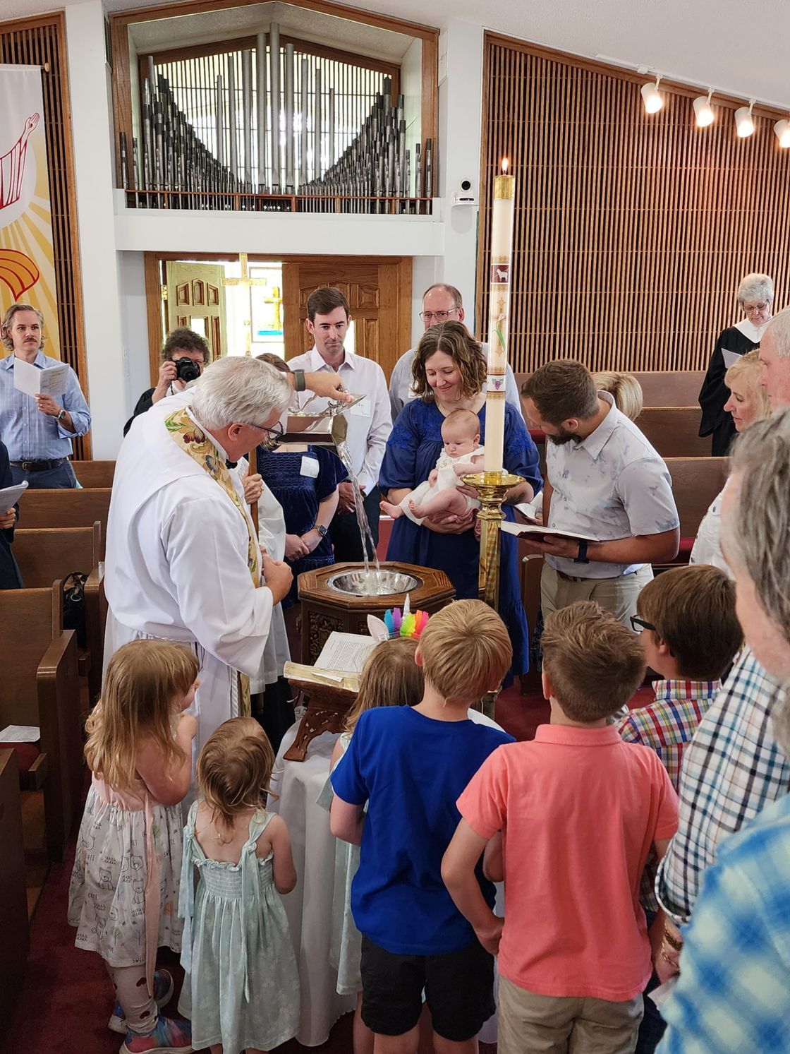 Baptism ceremony in a church. Priest pours water on baby held by a woman, surrounded by onlookers.