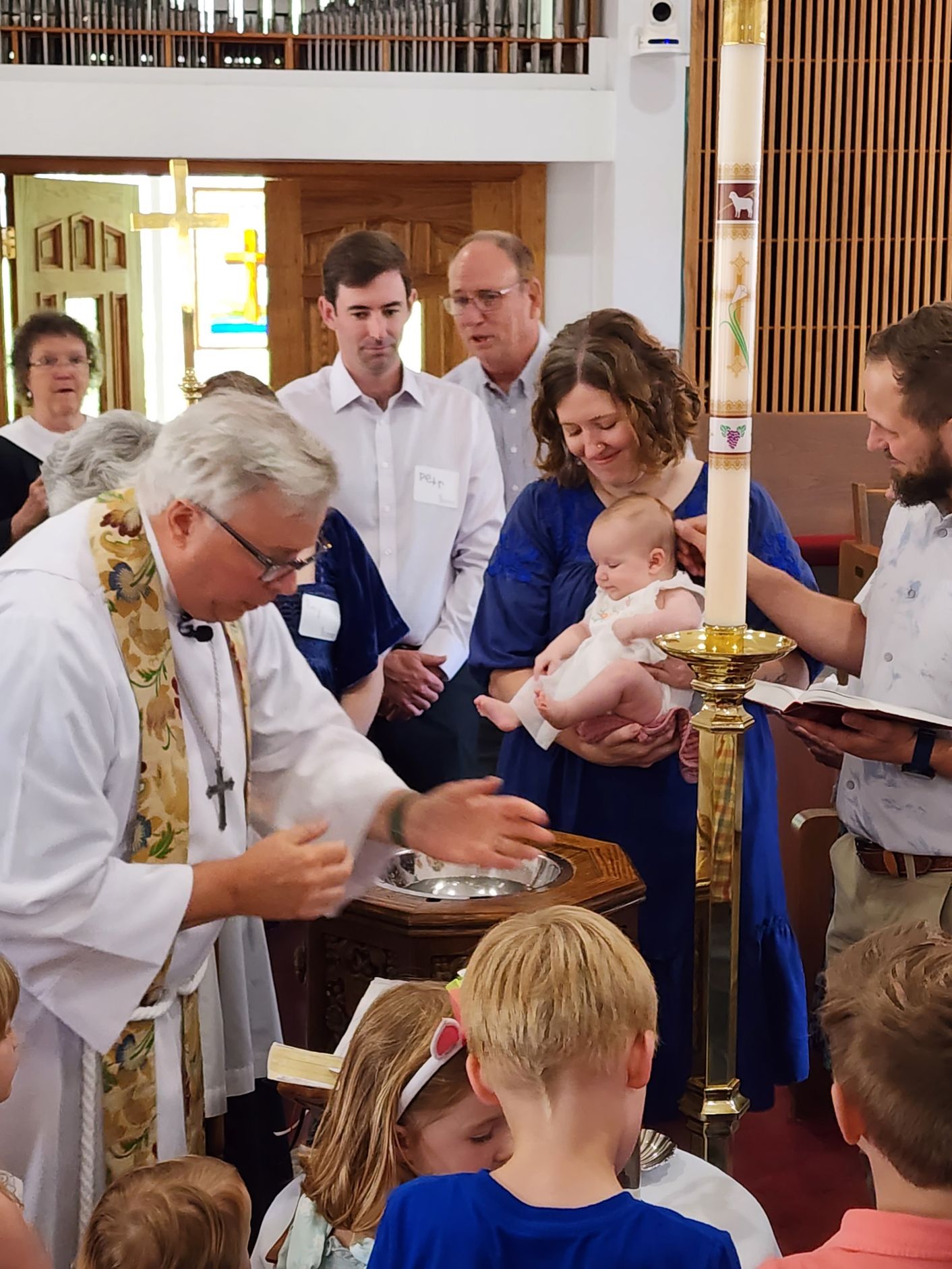 Priest baptizes a baby held by a woman in a church. People watch, including children.