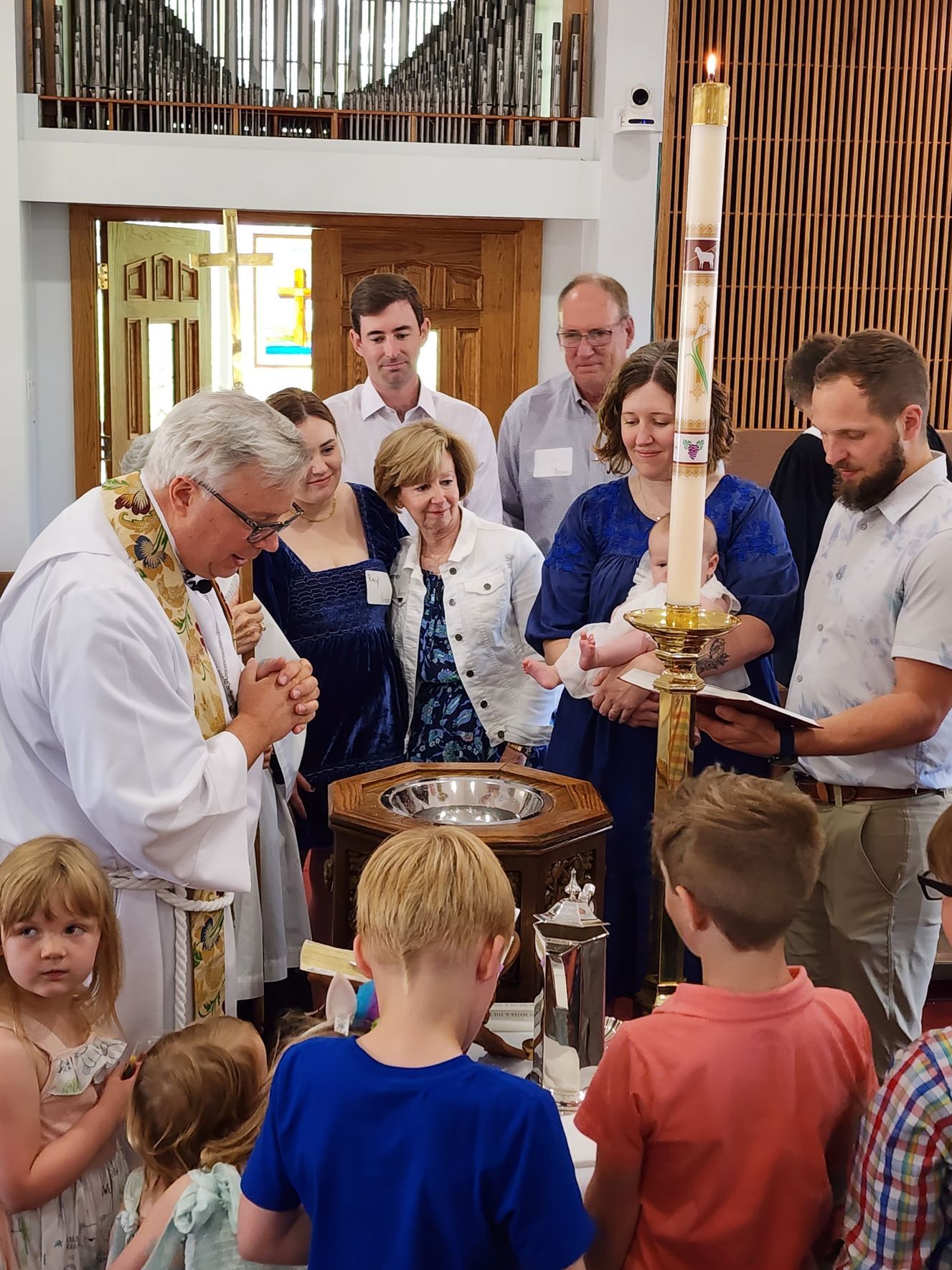 Priest baptizes a baby at a church baptismal font, surrounded by family and children.