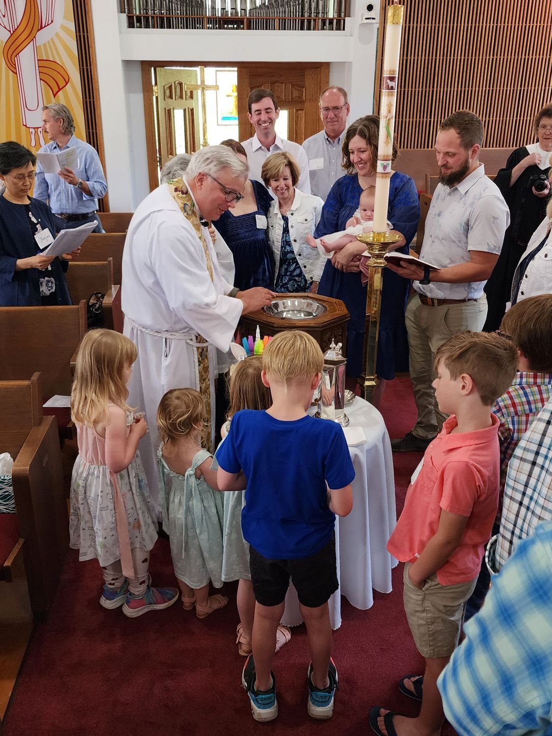 Priest baptizes a baby at a church. Several children and adults watch the ceremony.