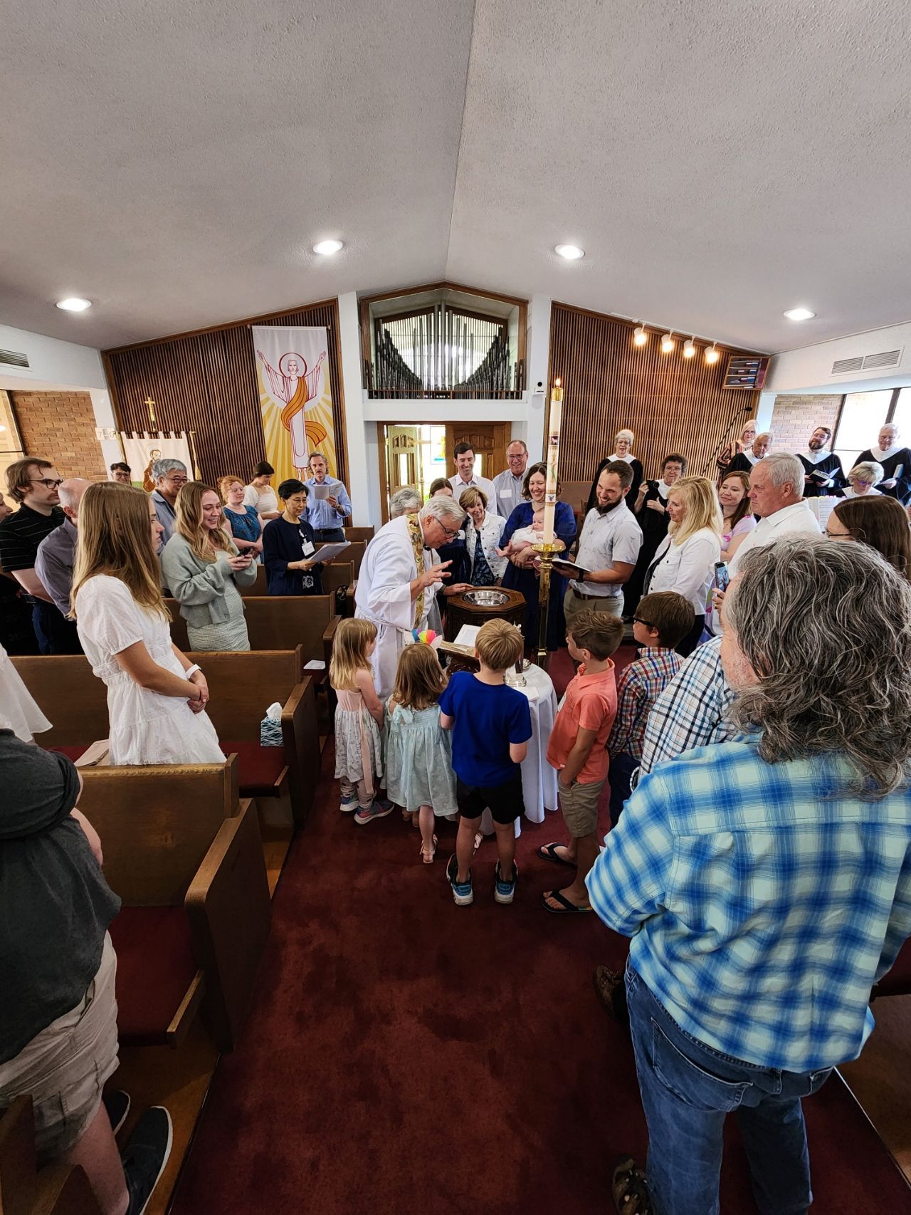 People in church for a baptism ceremony; a priest pours water over a child's head, surrounded by family and friends.