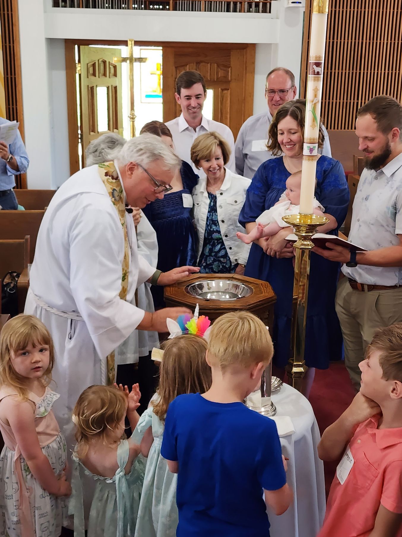 Baptism ceremony: Priest pouring water over a baby in a font, surrounded by family and children in a church.