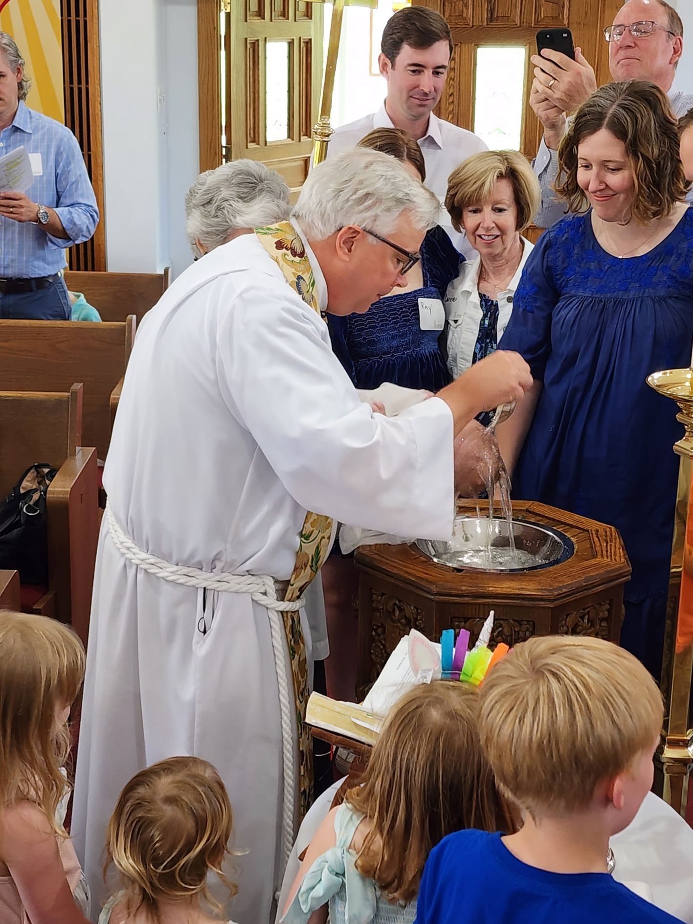 Priest baptizing a child in a church; people watch nearby.