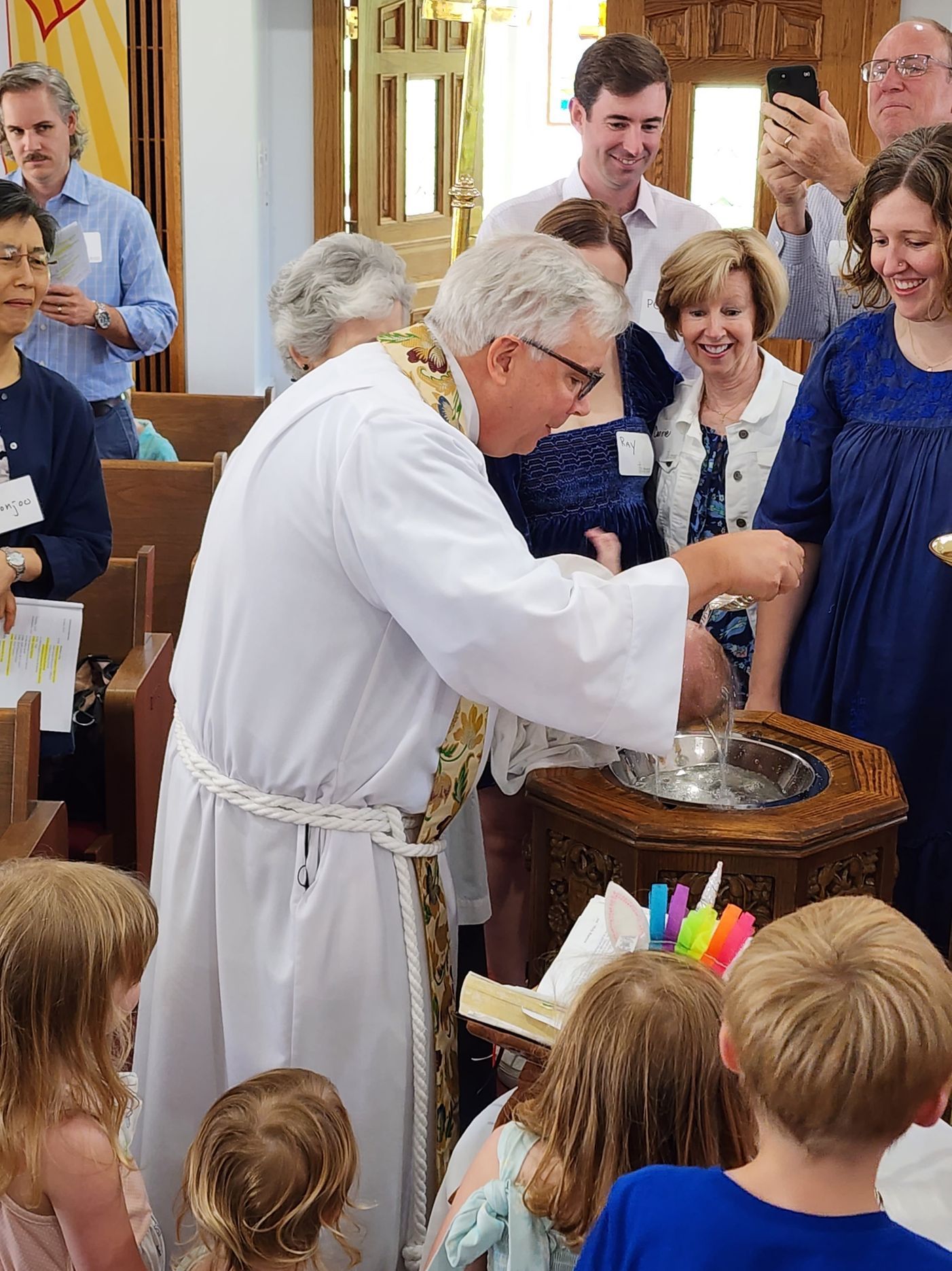 Clergyman baptizing a child in a church; people watch nearby.