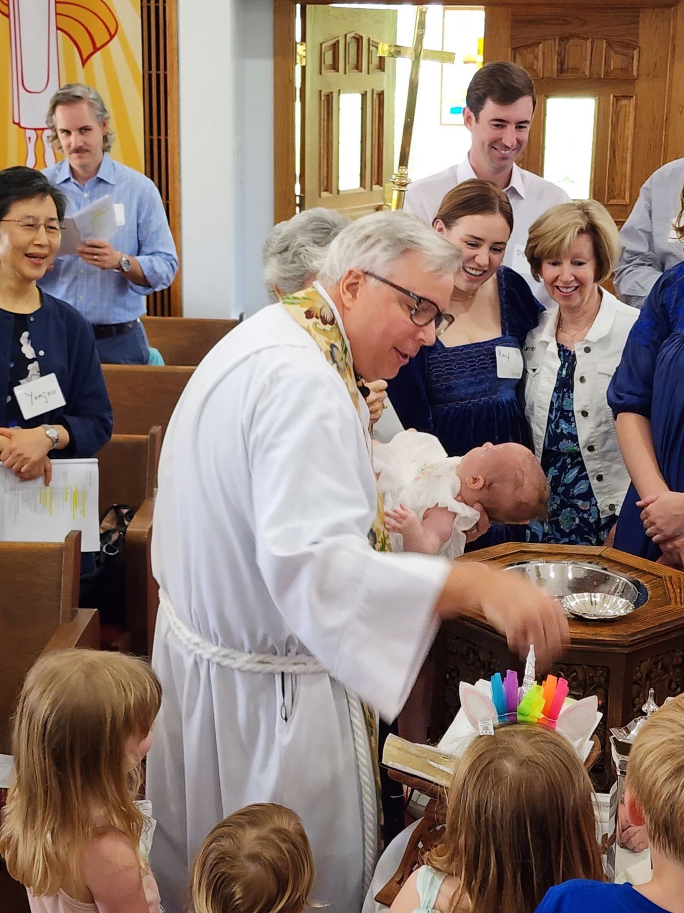 A priest baptizes a baby in a church, surrounded by family and children.