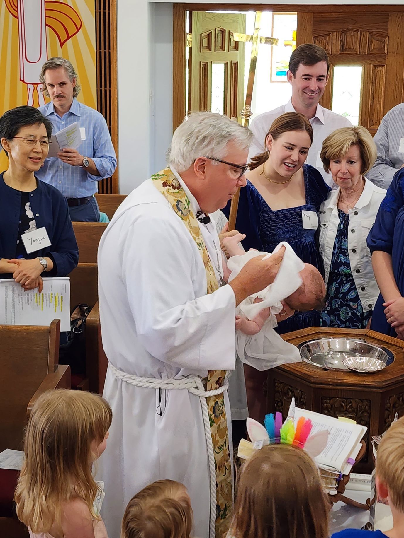 Priest baptizes a baby in a church. Family watches, smiling.