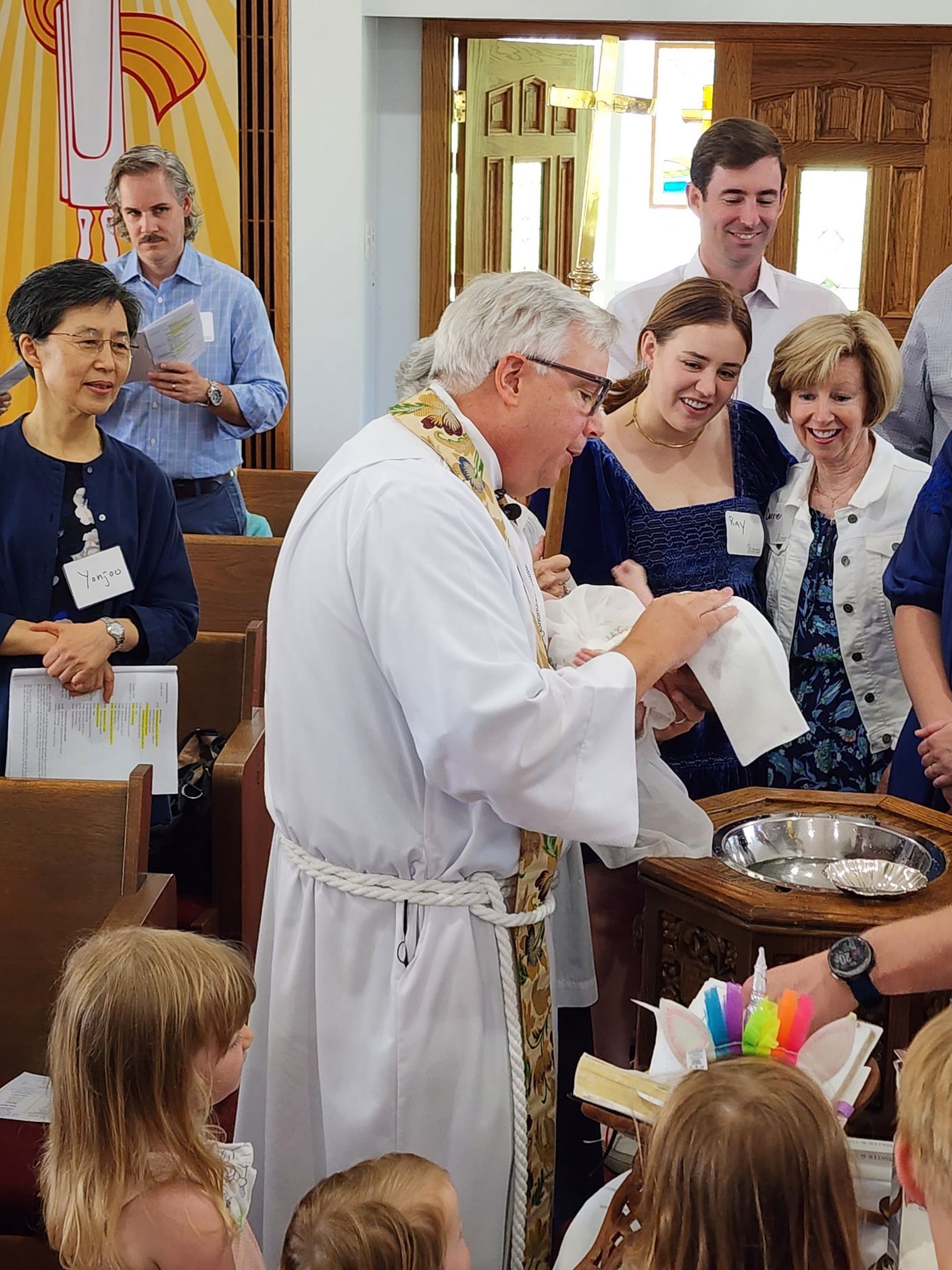 Priest baptizing a baby in a church, surrounded by family and attendees; a baptismal font is visible.
