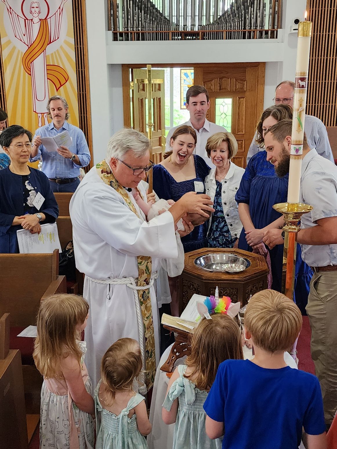 Clergy baptizes a baby in a church, surrounded by family and children, with joy and anticipation.