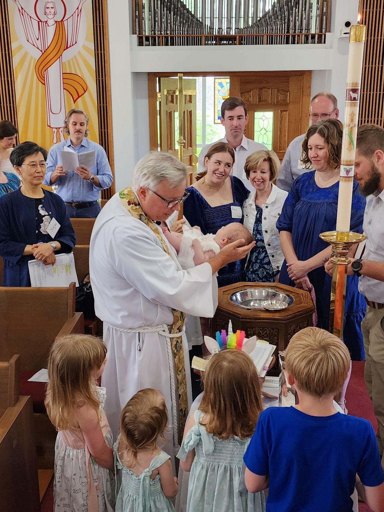 Clergy baptizing a baby in a church, surrounded by family and children.