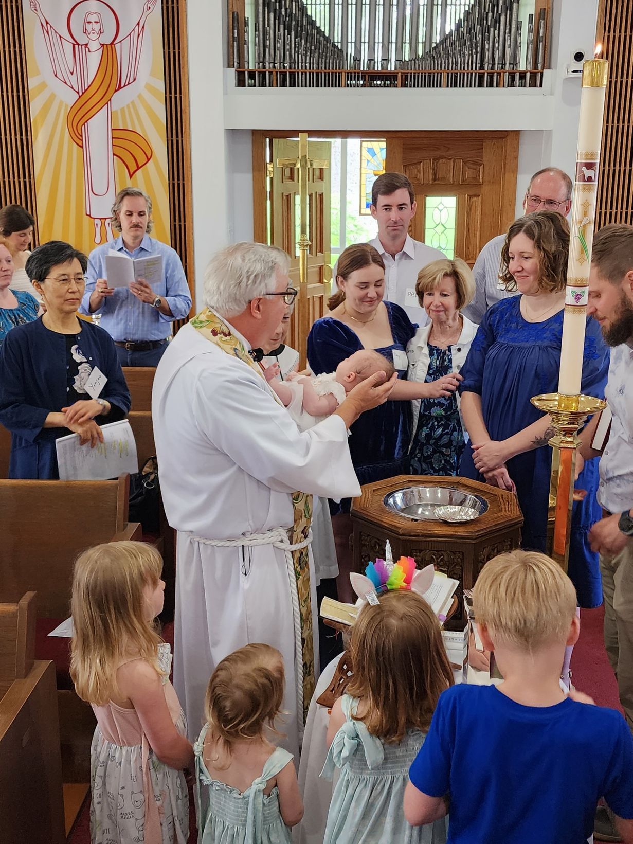 Priest baptizes a baby at a church. Family watches near the baptismal font, candles lit.