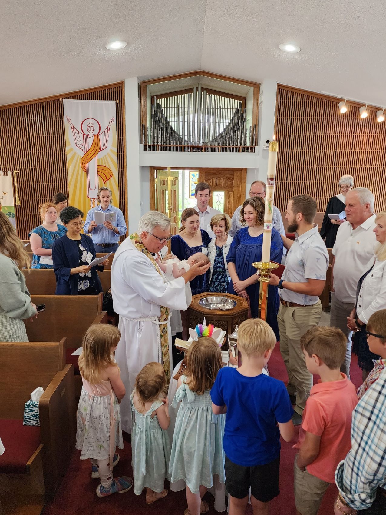 Baptism ceremony in a church; priest pouring water over an infant held by a parent. Family and guests watch.