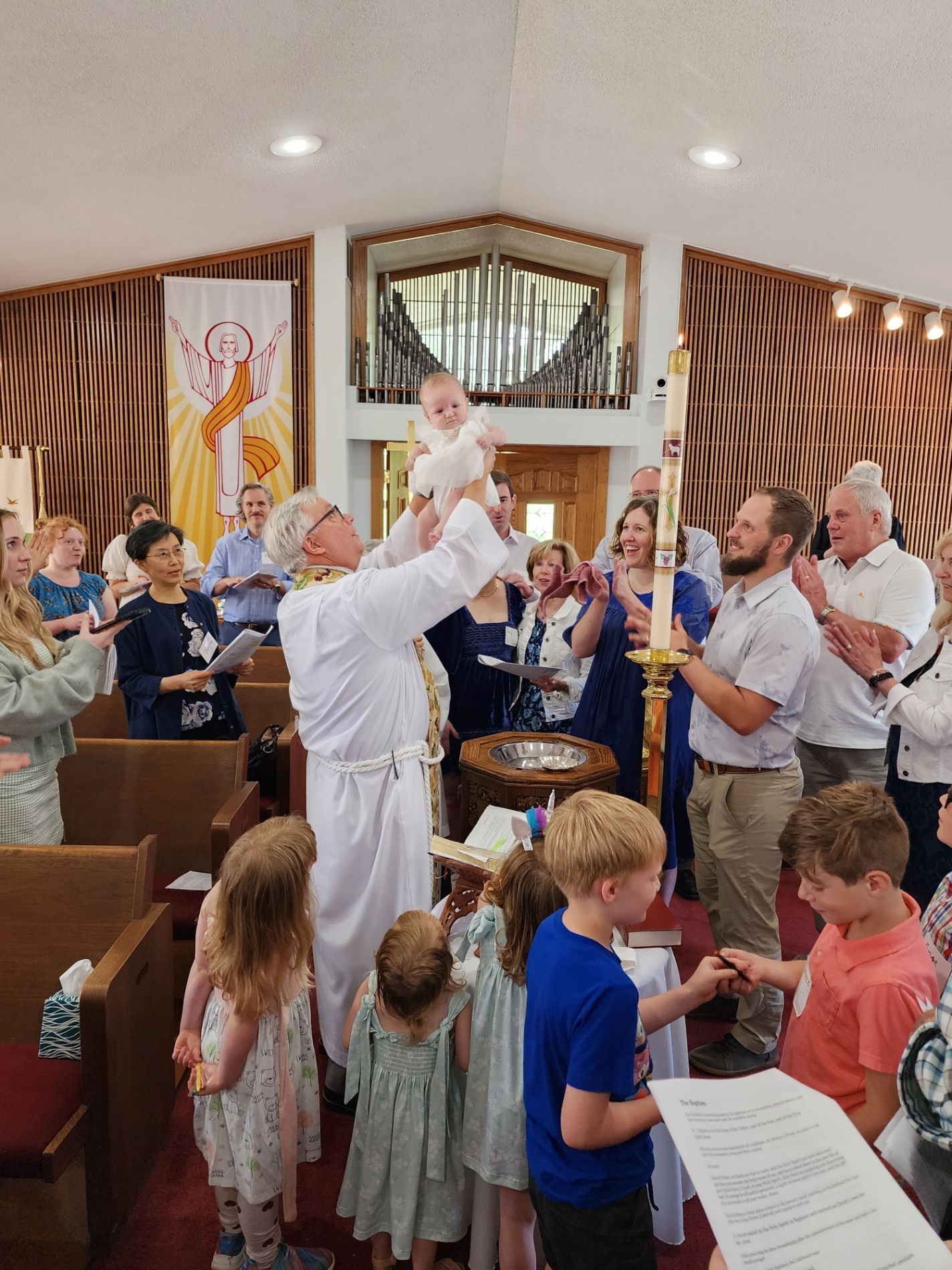 Priest holds baby aloft during baptism ceremony at church. Family and friends watch.
