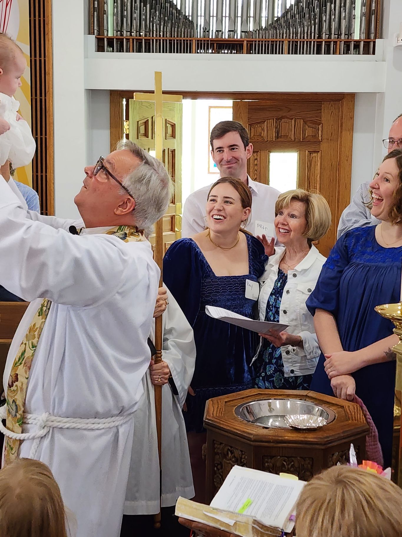 Priest baptizes a baby in a church; family watches and smiles.