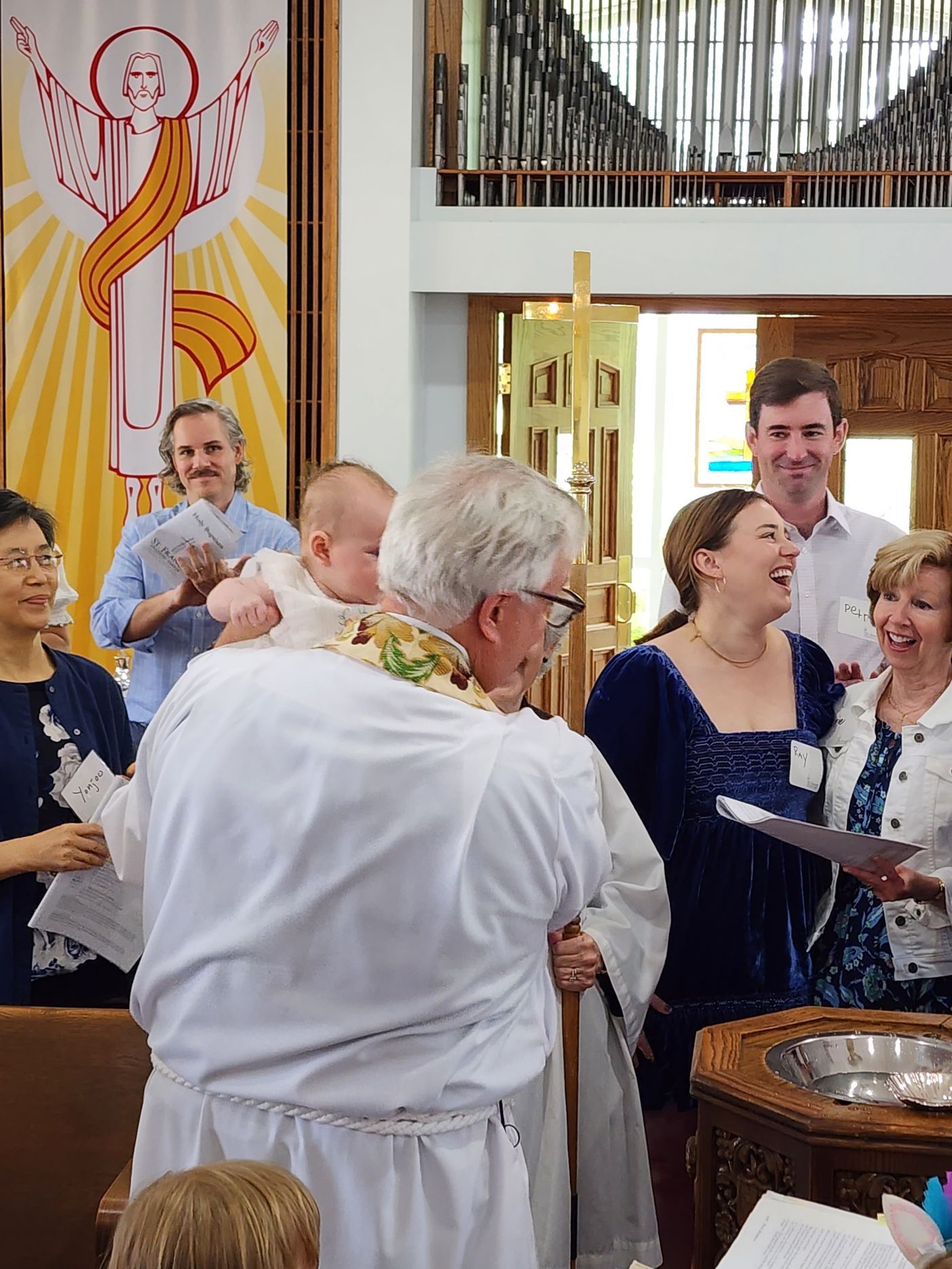 A priest baptizes a baby in a church, surrounded by smiling attendees and a banner.