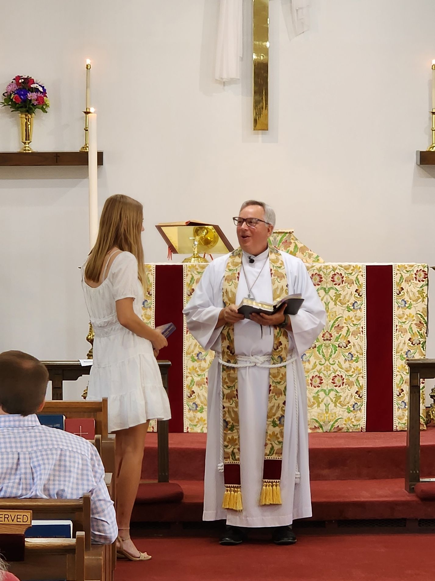Clergyman speaking to a woman in a white dress at a church altar; candles and floral arrangement in the background.