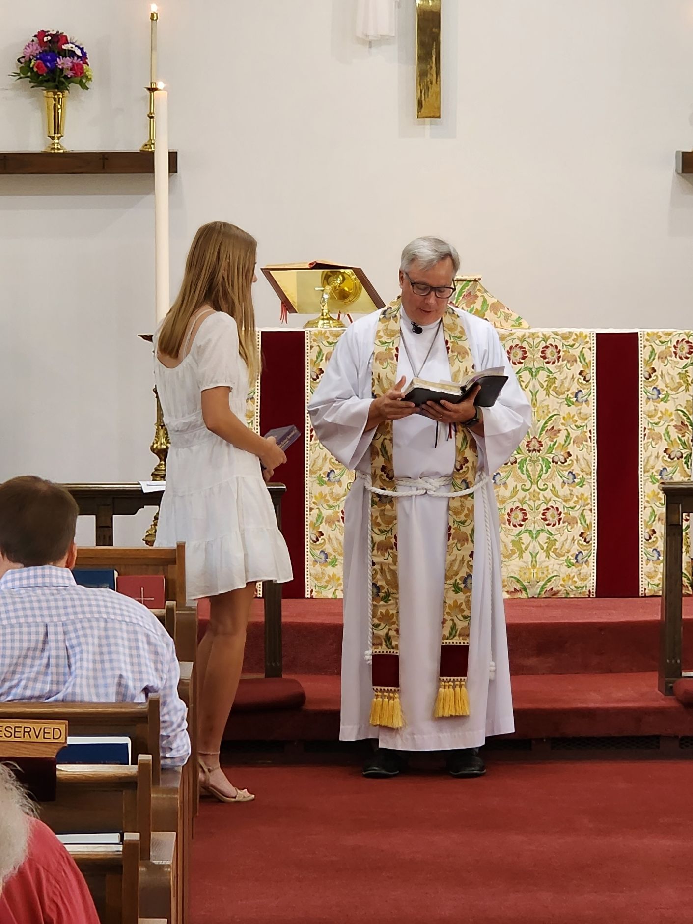 Clergyman in white robes reads from a book, addressing a woman in white dress, standing inside a church.
