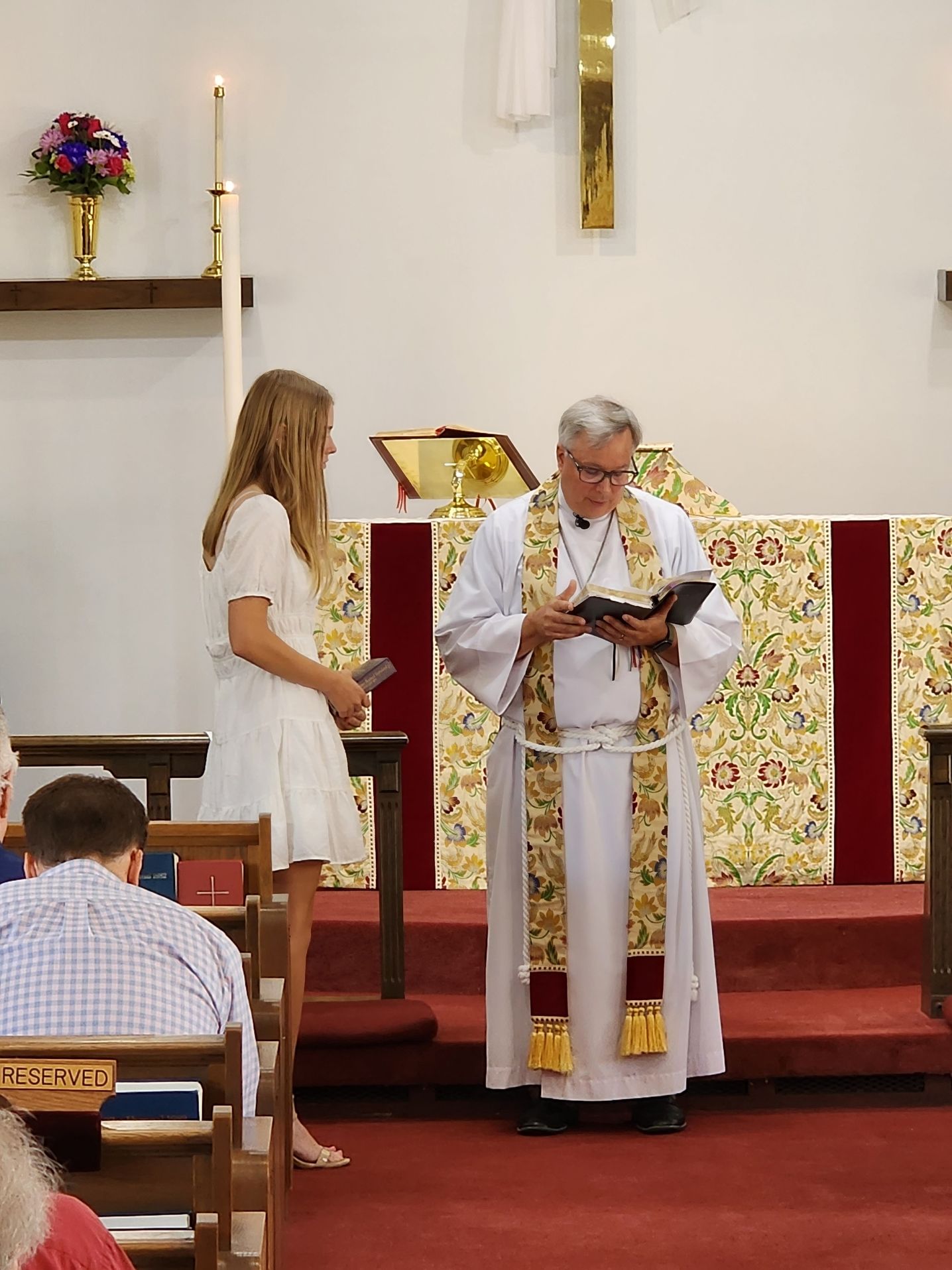 A person in white dress stands beside a robed officiant reading from a book in a church.