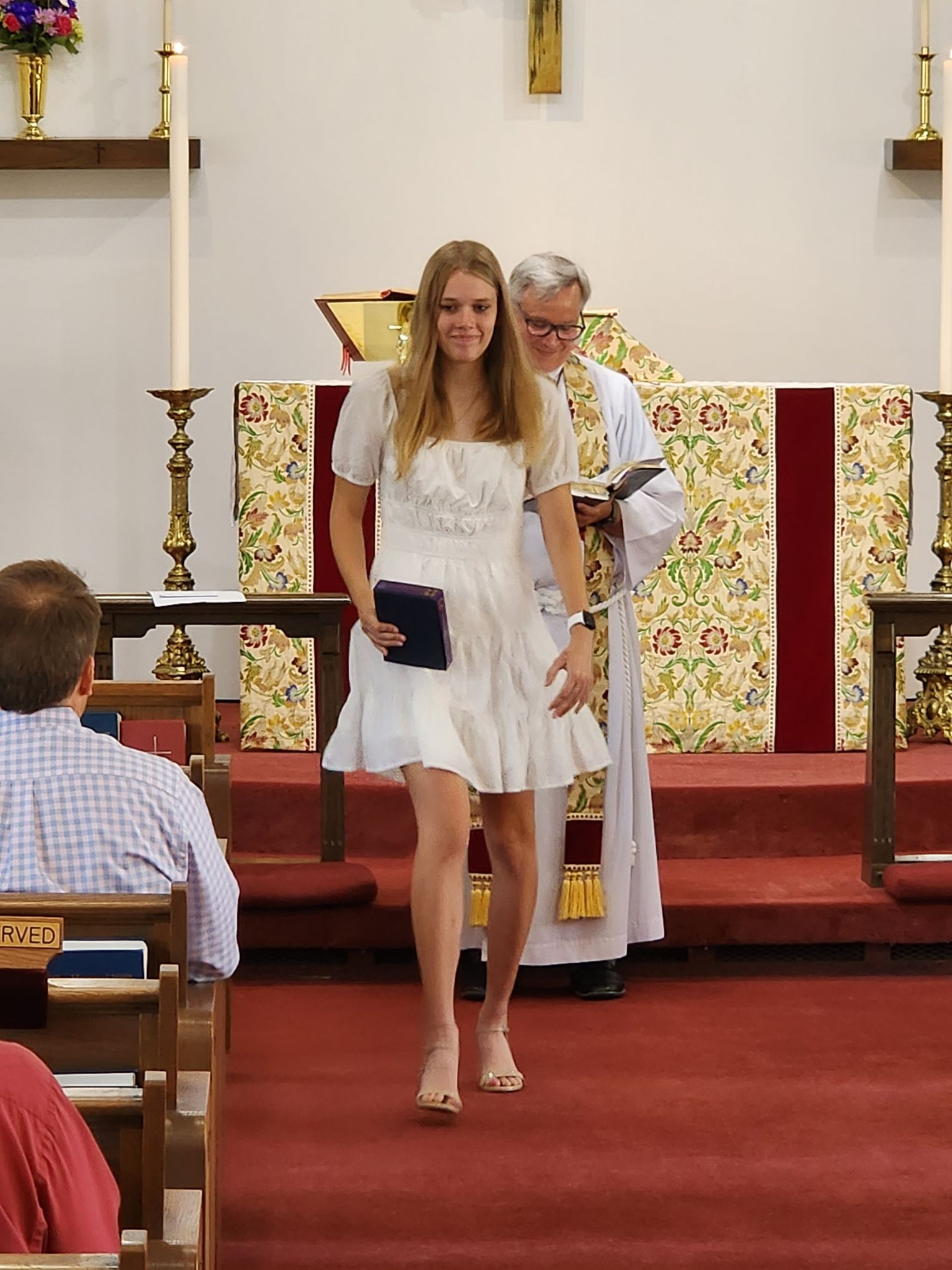 Woman in white dress walks down church aisle, followed by a person in a robe.