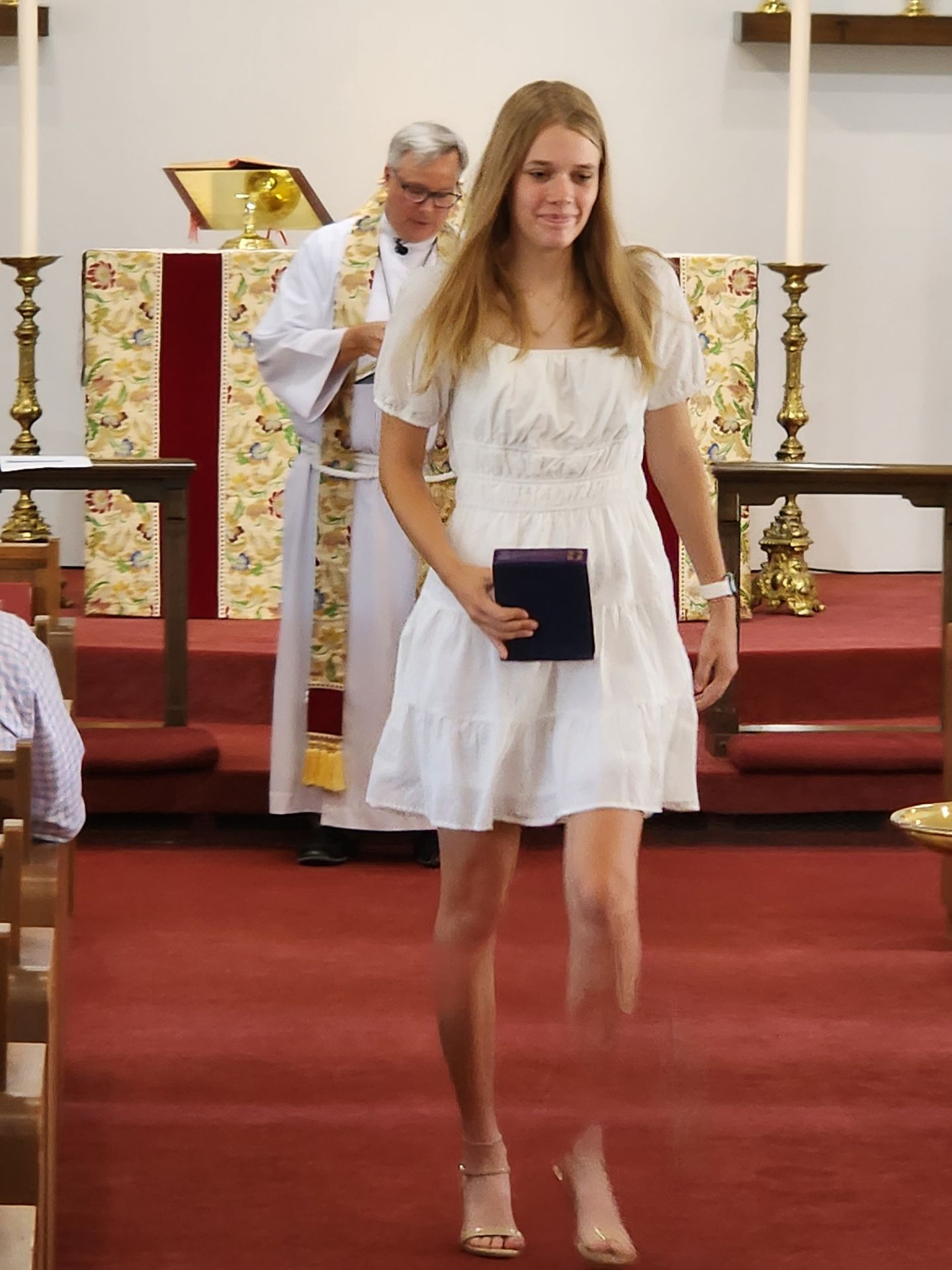 Woman in white dress, holding a book, walks forward in a church, with a priest and altar in the background.