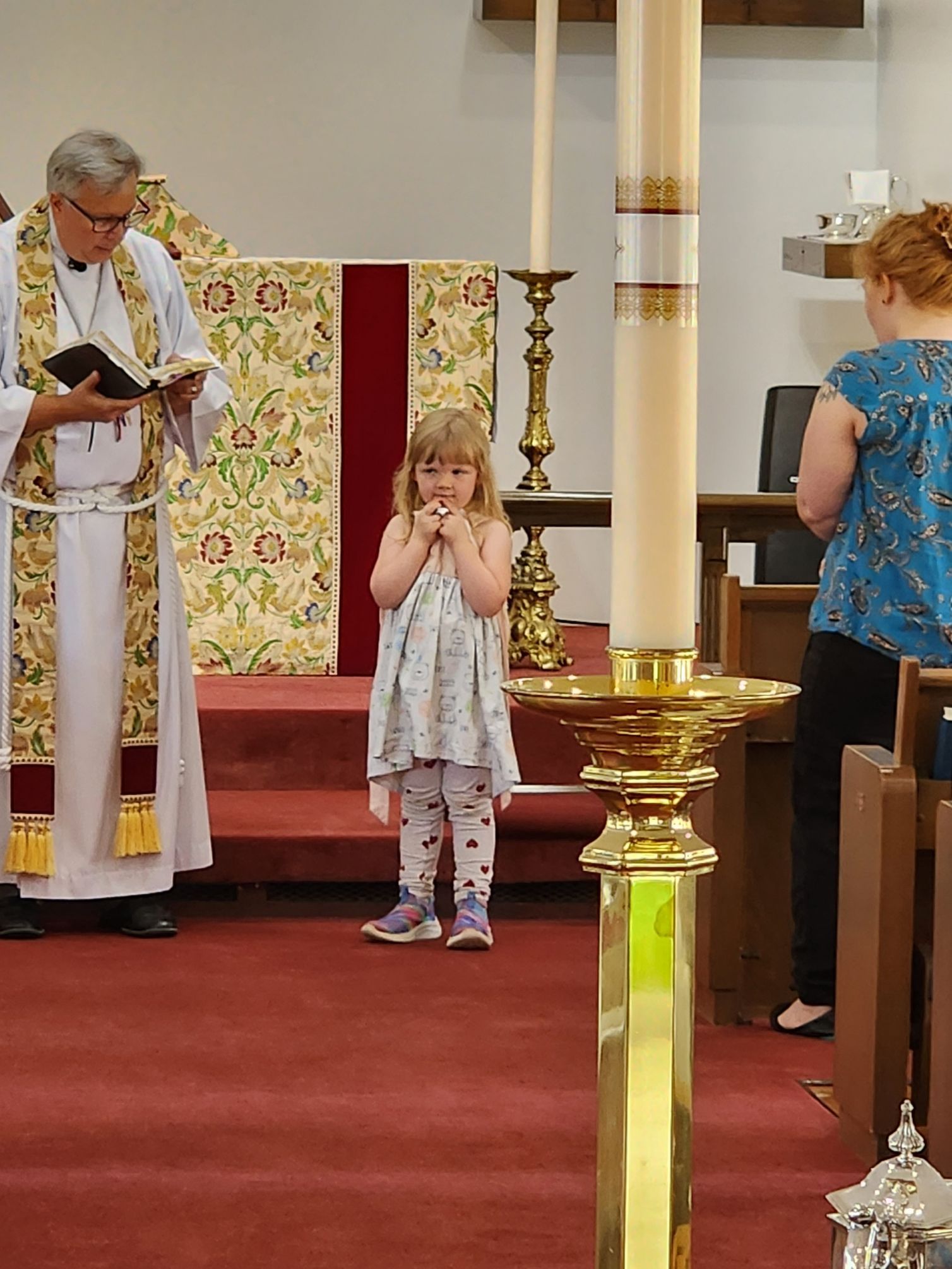 A child standing at an altar, hands clasped, in a church, with a priest and another person looking on.