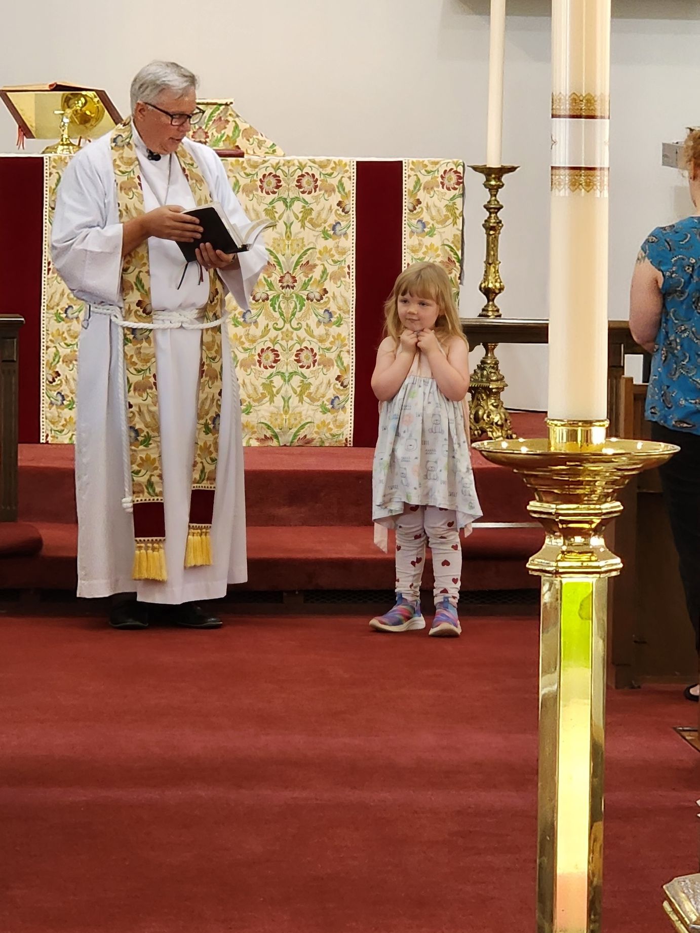 Clergyman in robes reads to a young child at a church altar. The child has hands clasped and is looking up.