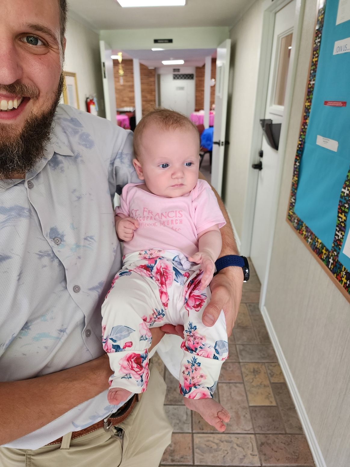 Man holding a baby in a hallway; baby wears pink top and floral pants.
