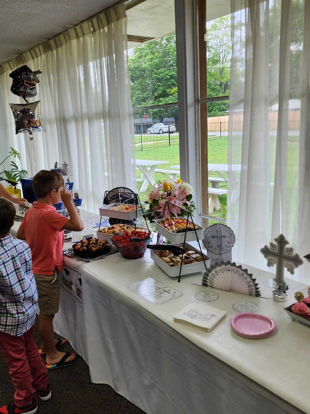 Buffet table with snacks, flowers, and decorations inside a building, children standing nearby.