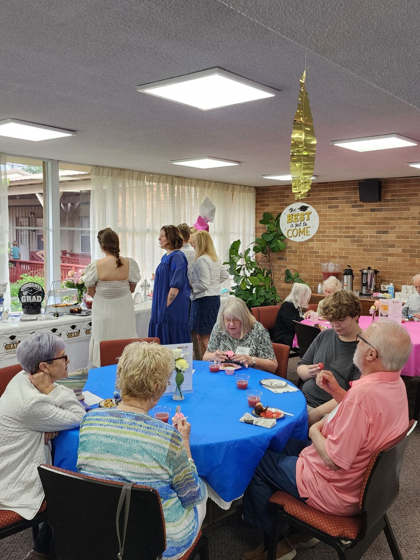 People seated at tables, socializing in a room, with a window and decorations.