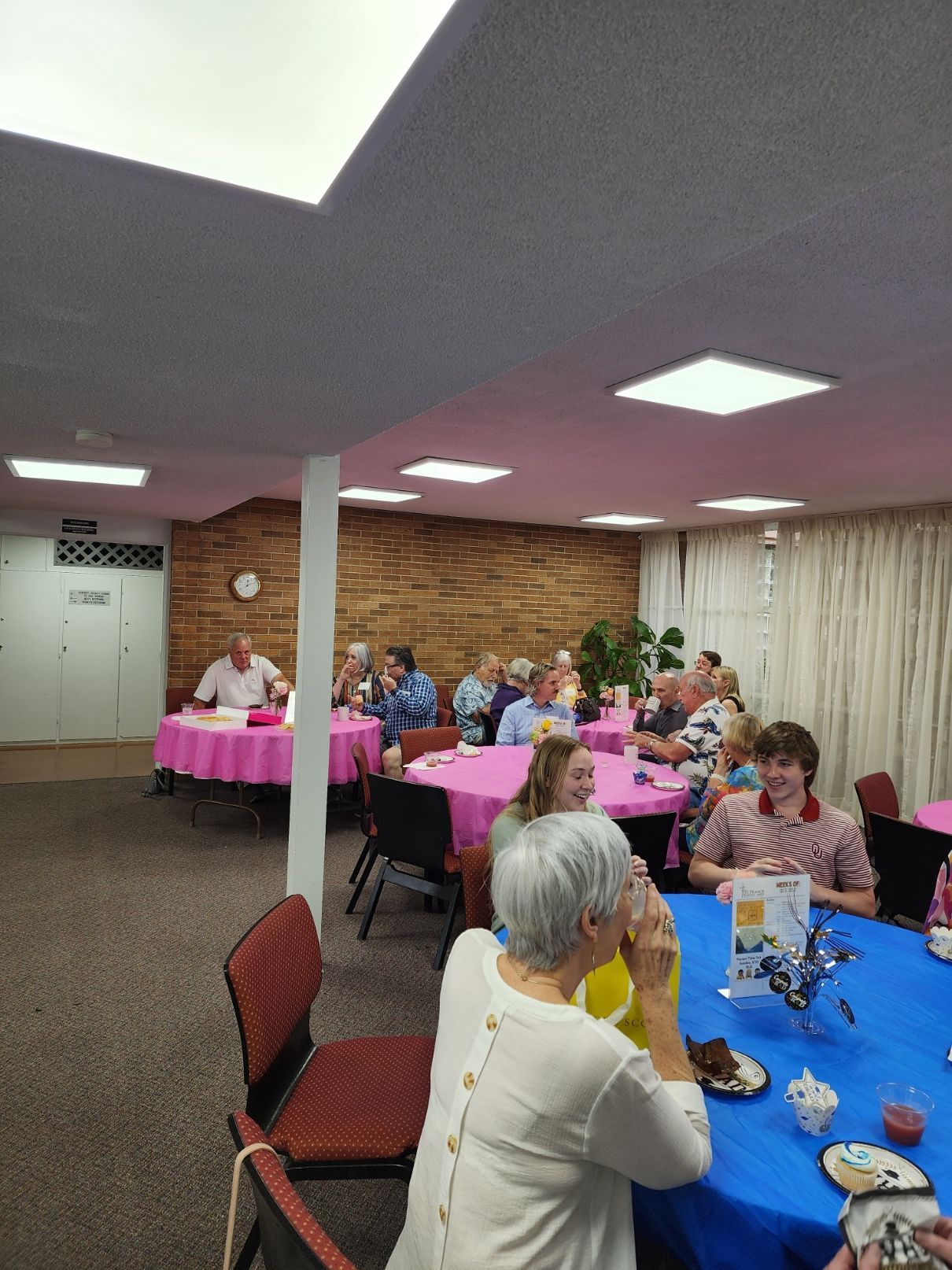 People seated at tables with pink and blue tablecloths in a room, some are eating or talking.
