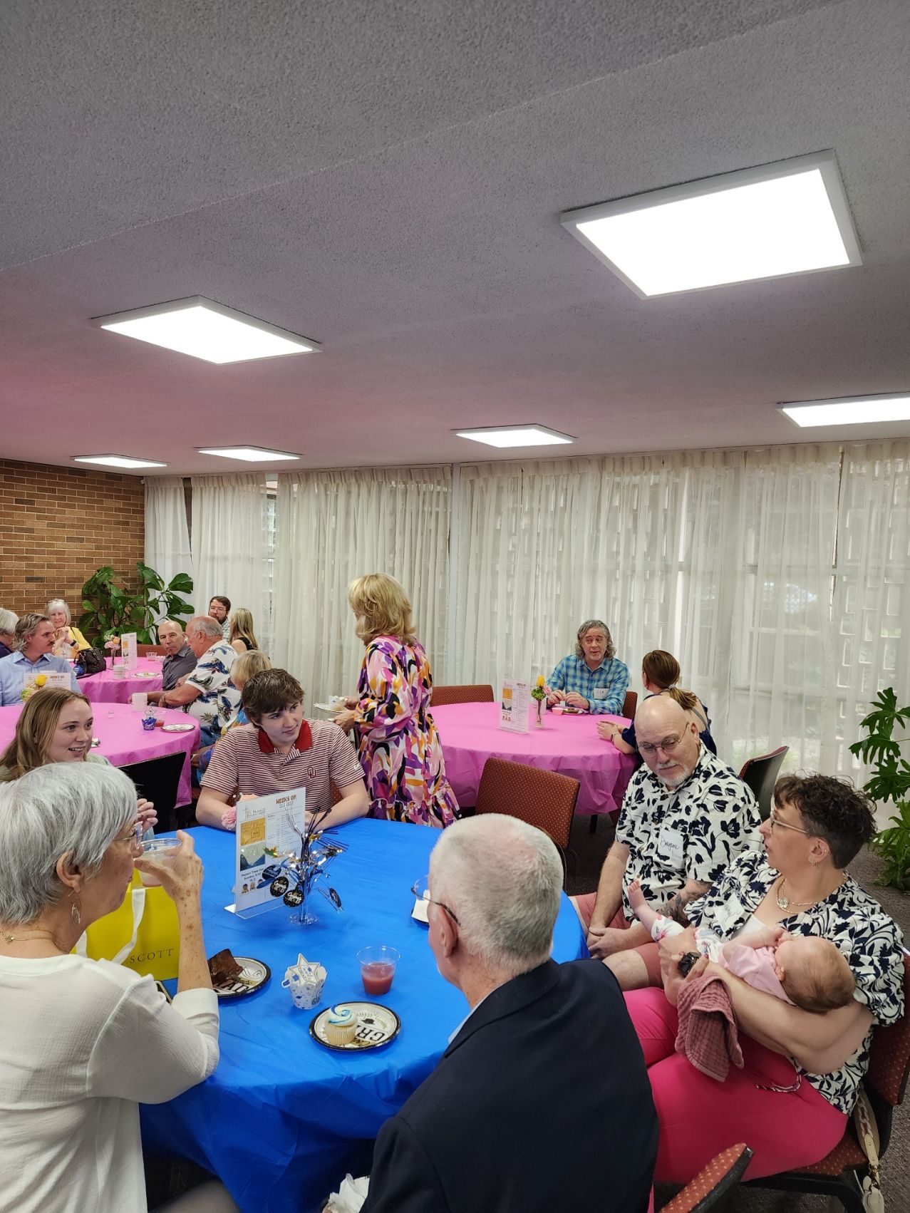 People seated at tables with blue and pink tablecloths, in a brightly lit room; one woman is holding a baby.