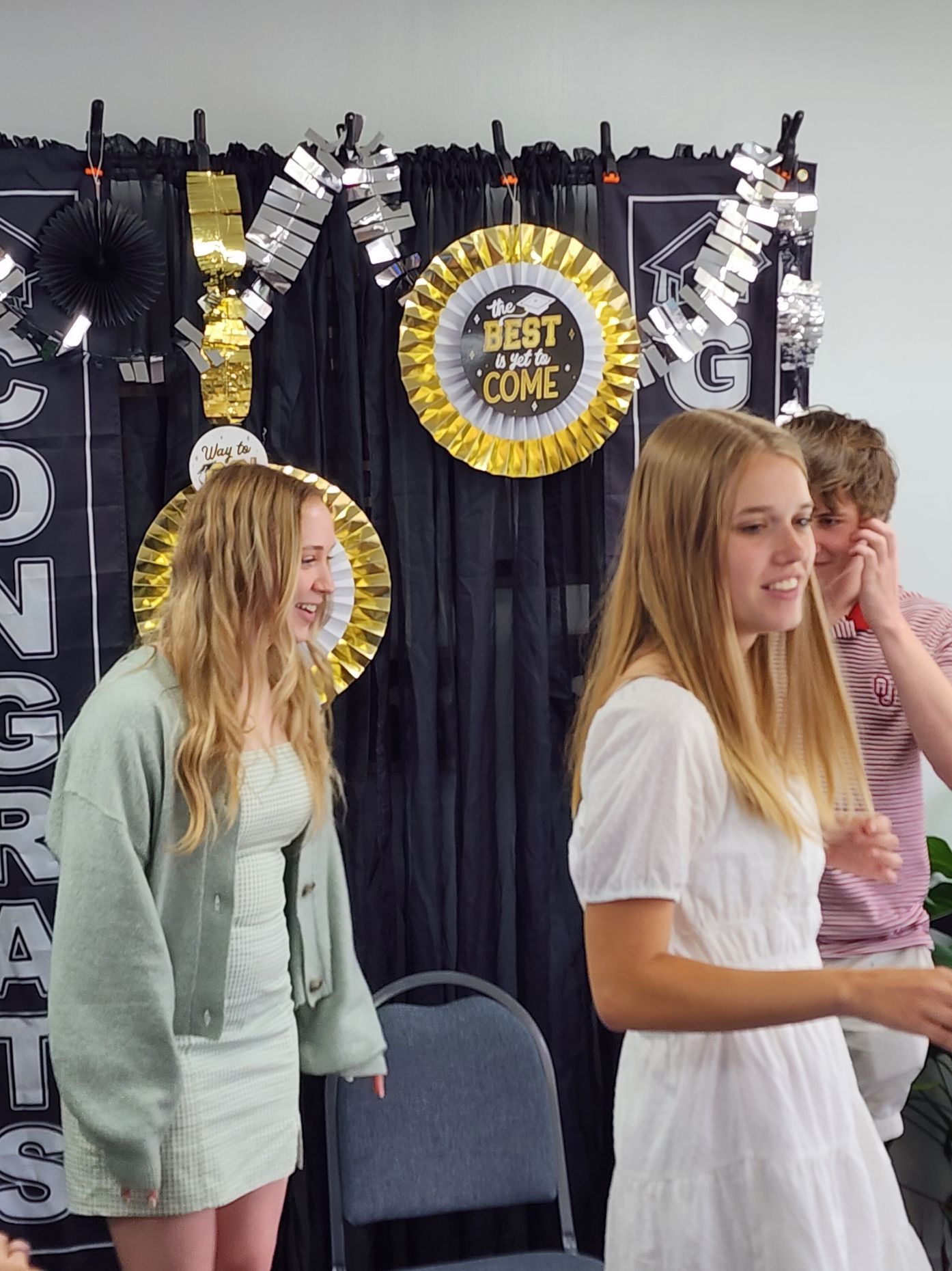 Two young women and a young man in front of a graduation backdrop. One woman wears a white dress. The other a light green sweater.
