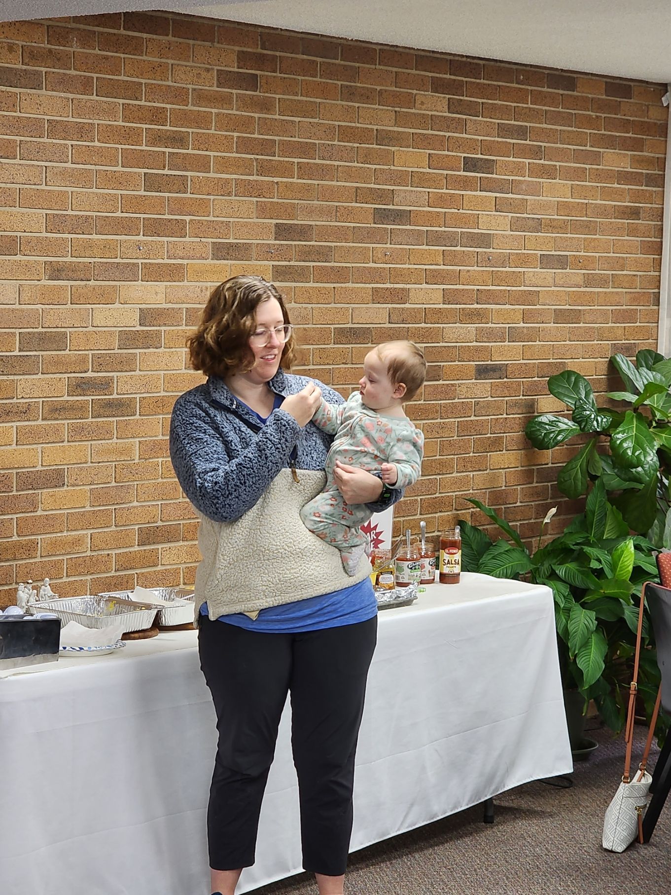 Woman holding a baby, smiling, near a table with food, in front of a brick wall.