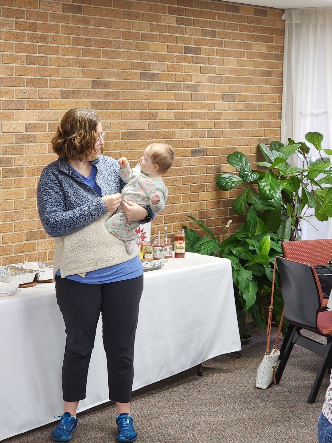 Woman holding a baby, smiling at each other, near a brick wall and a table with items on it.