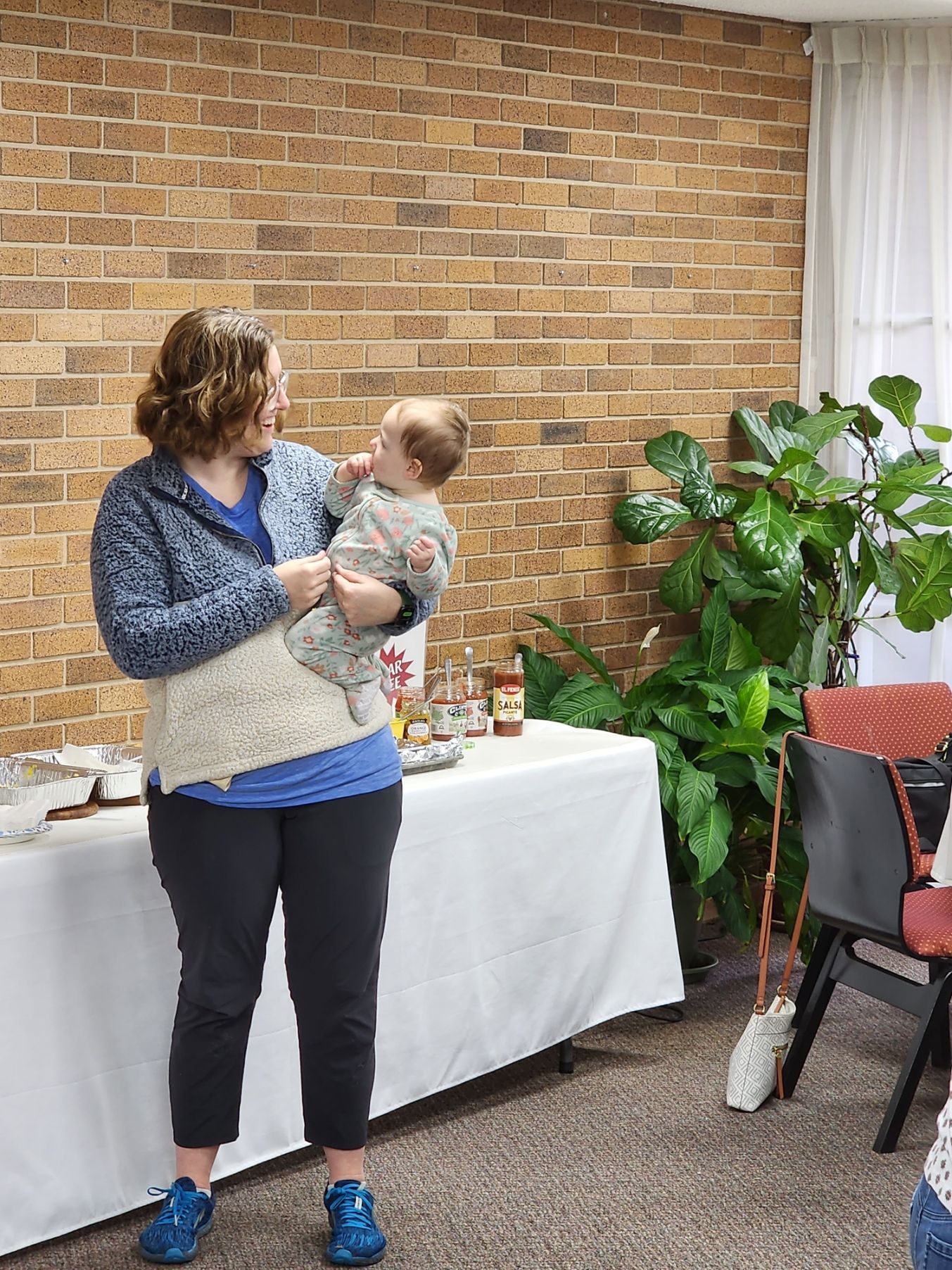 Woman holding a baby smiles, standing by a table with food, in front of a brick wall and plants.