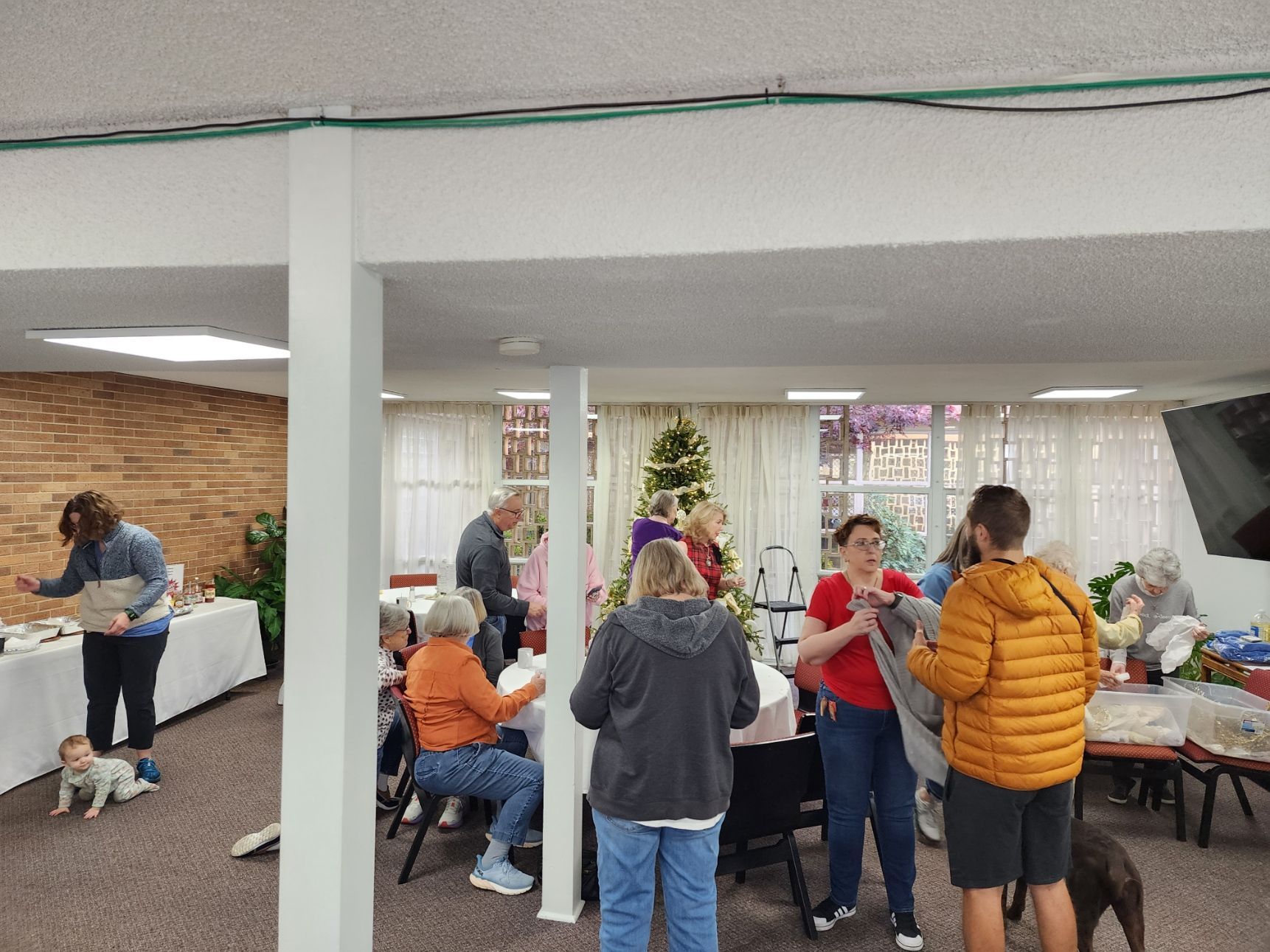 People gather in a room with tables and a Christmas tree. Some are looking at items, some are talking.