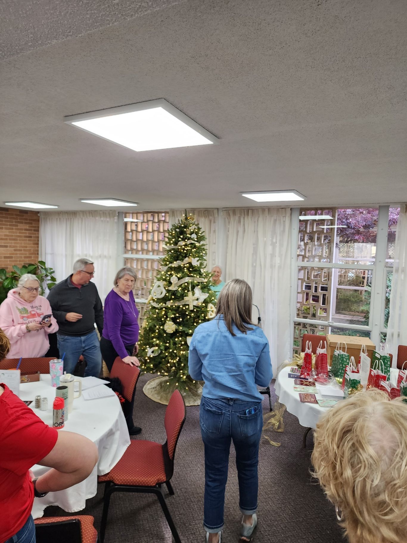 People in a room with a Christmas tree. One woman speaks, others look on. Tables with gifts are visible.