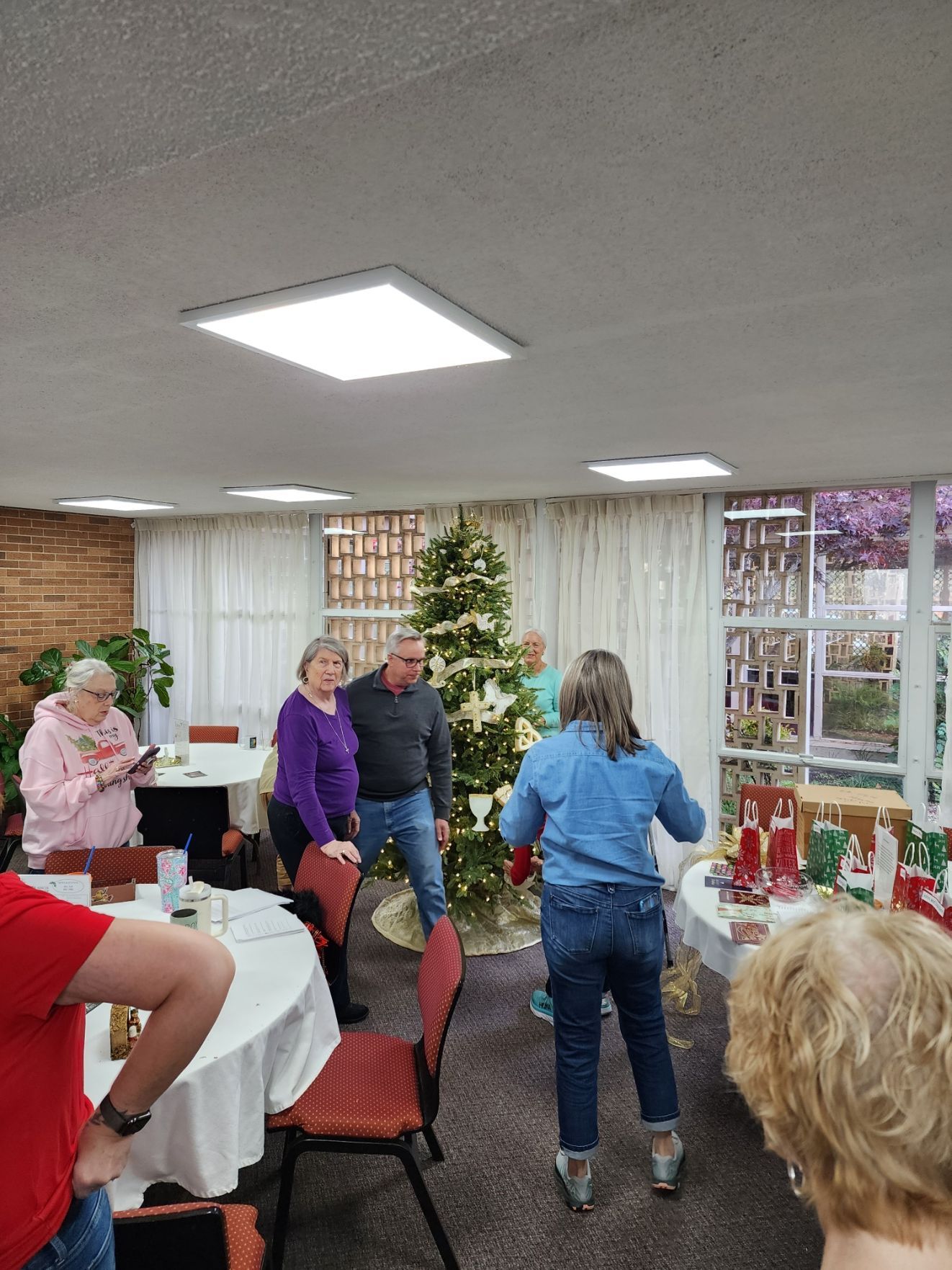 People around a Christmas tree, some looking at it, in a room with a window and a round table.