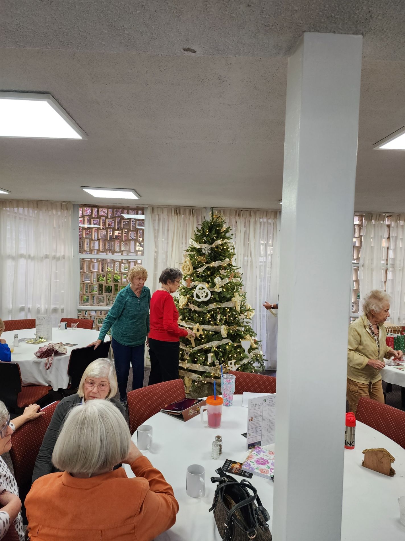People decorate a Christmas tree in a room with tables. One person stands near a white pillar.