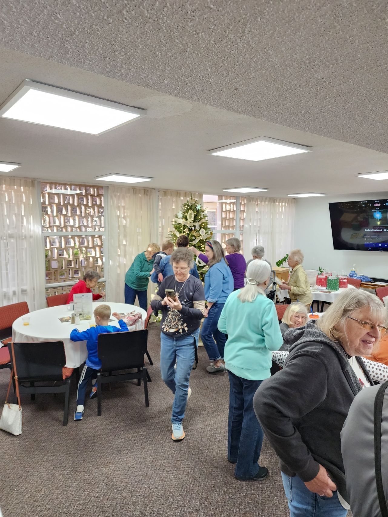 People gathered indoors around tables and a Christmas tree; some are talking and looking at presents.