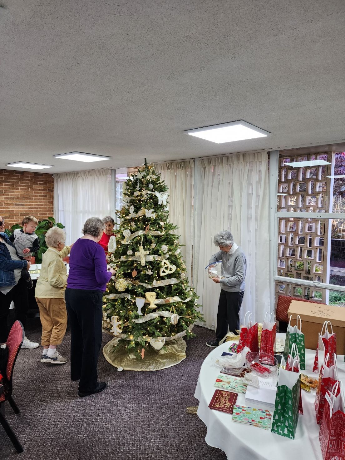 People decorating a Christmas tree indoors, surrounded by gifts on a table.