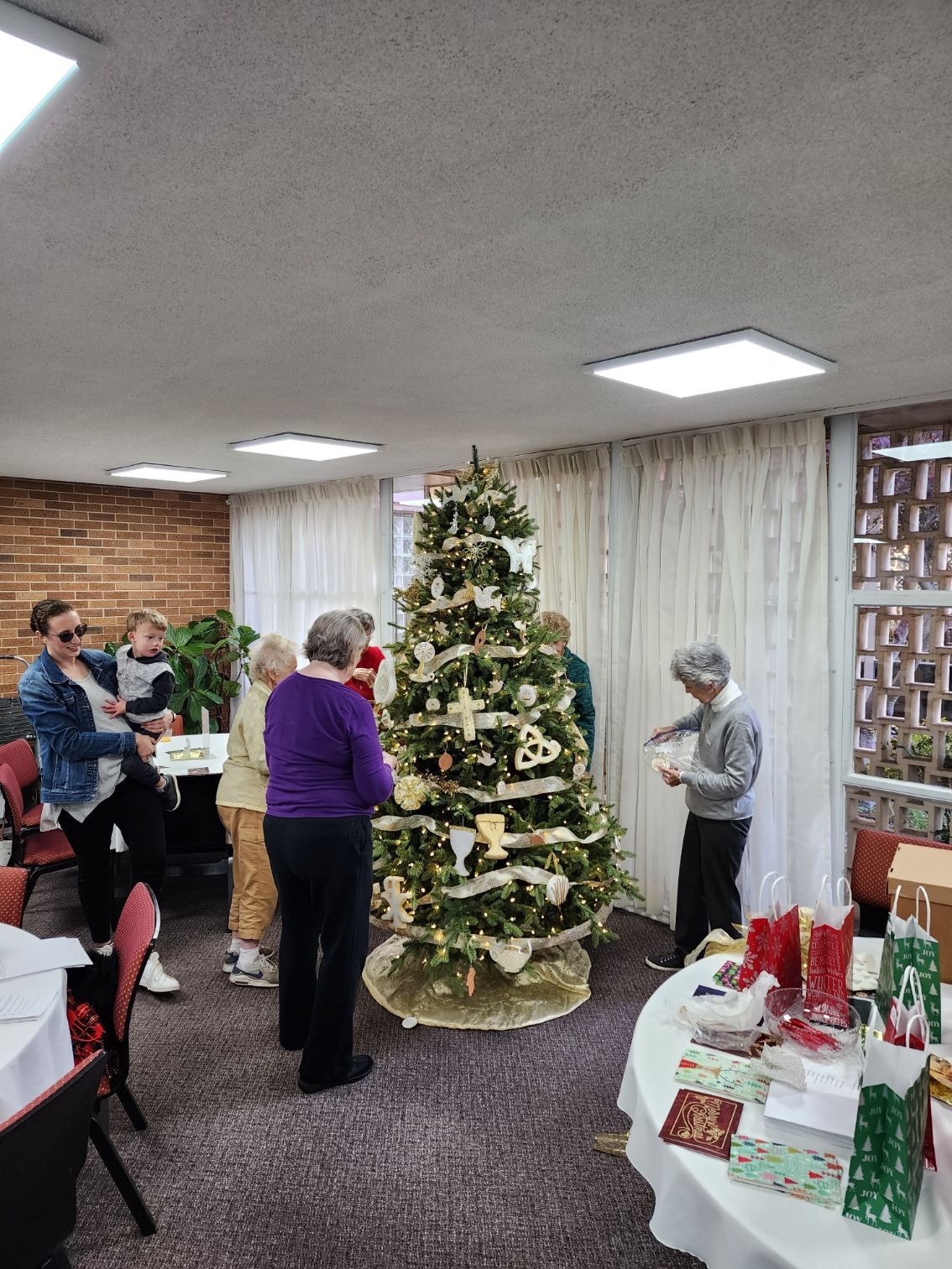 People decorating a Christmas tree in a room with a table of gifts; some are holding decorations, some are looking on.