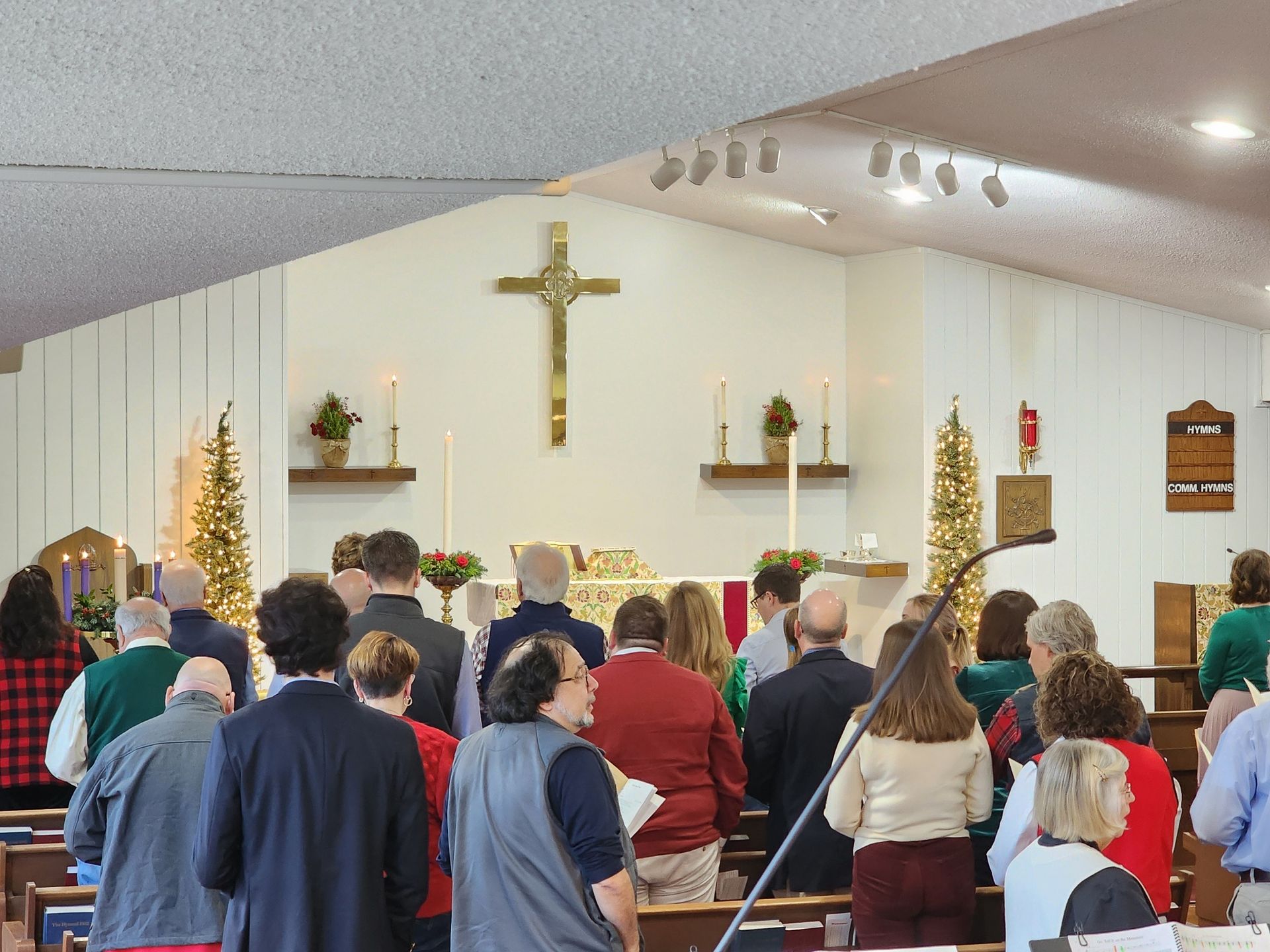 People standing in a church, facing altar with cross. Some hold songbooks. Christmas decorations are present.