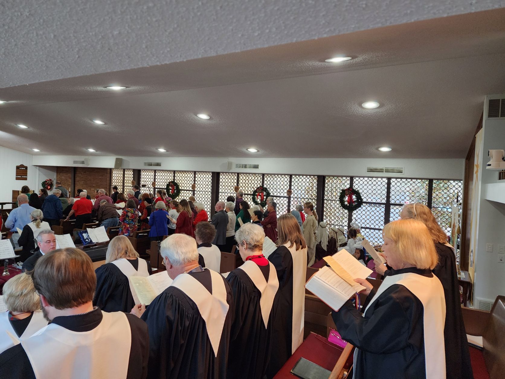Choir in robes singing during a service. Attendees in background, windows with wreaths.