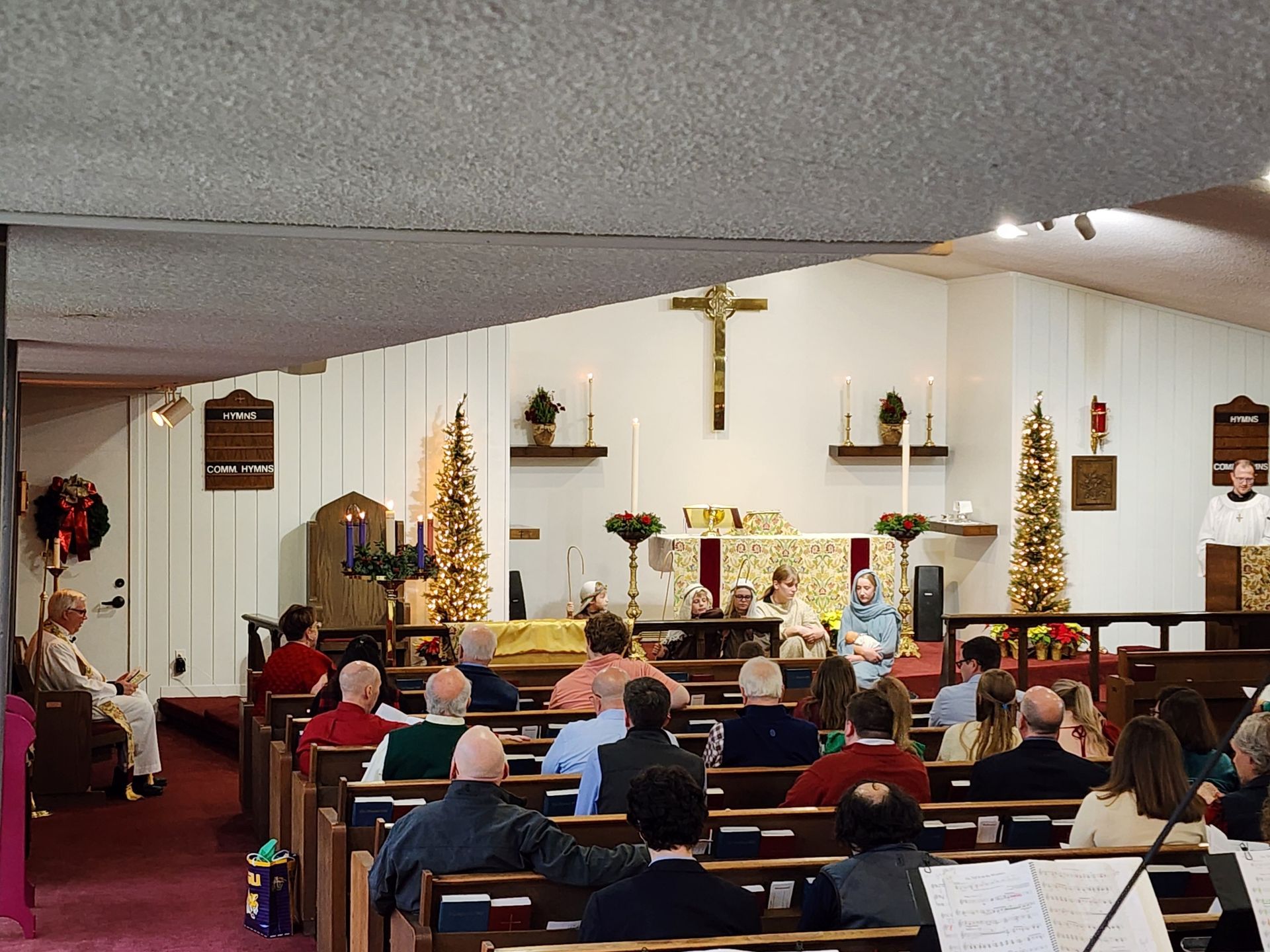 Church service in progress: people seated, altar with cross, decorated with candles and Christmas trees.