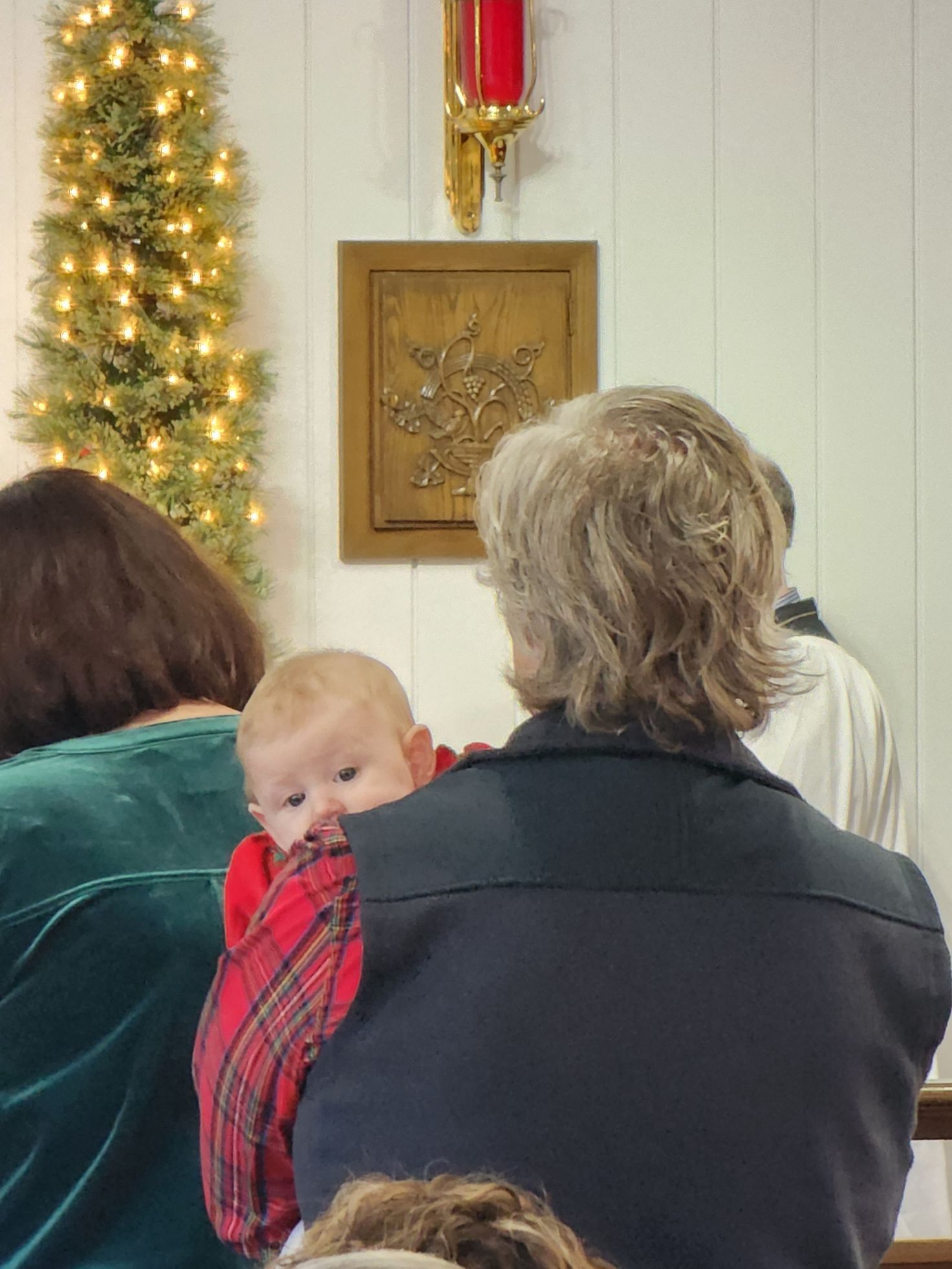 Person holds a baby wearing a red plaid outfit inside a decorated room with a Christmas tree.