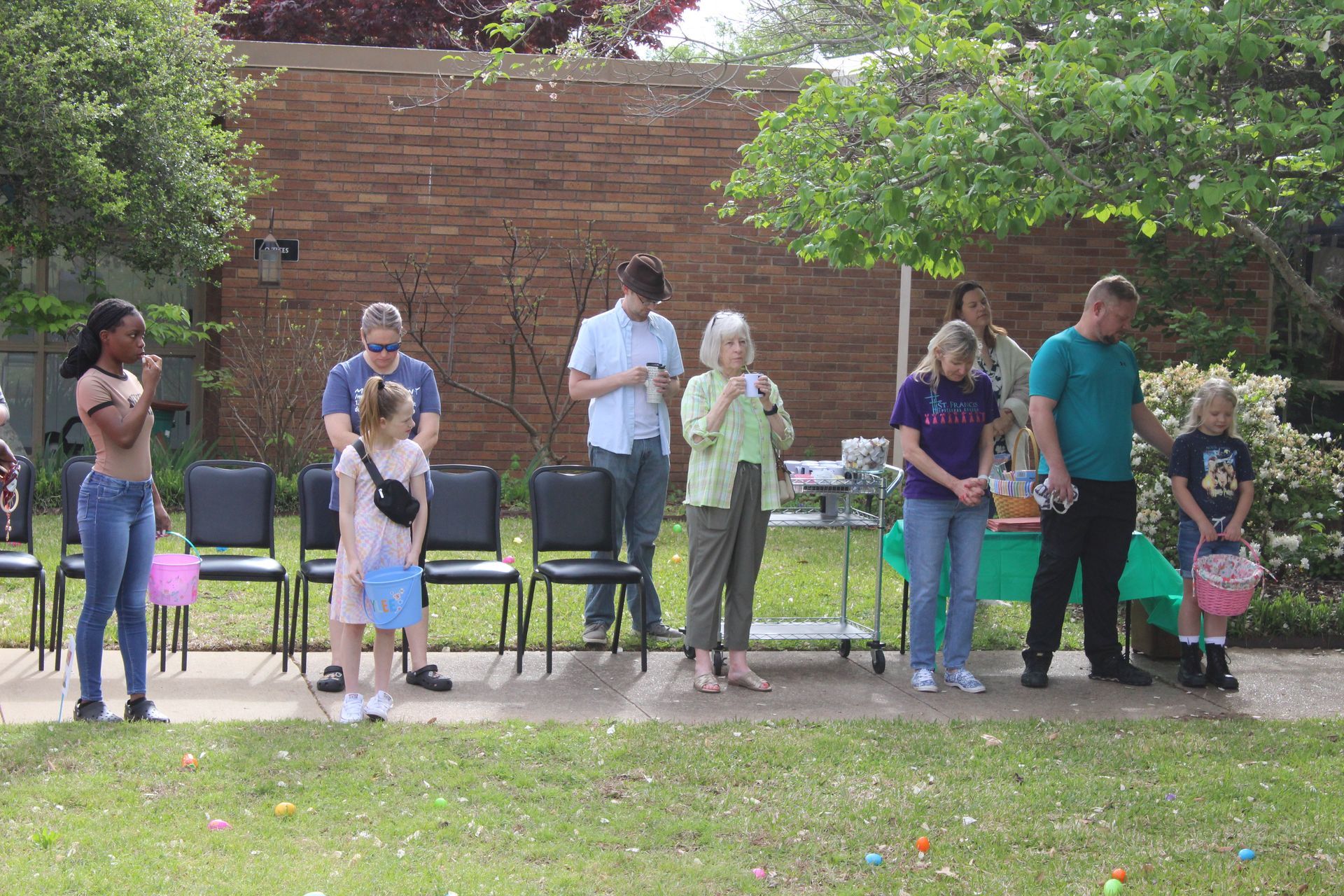 People at an outdoor event with Easter baskets, looking towards a grassy area with scattered eggs; near a brick wall and trees.