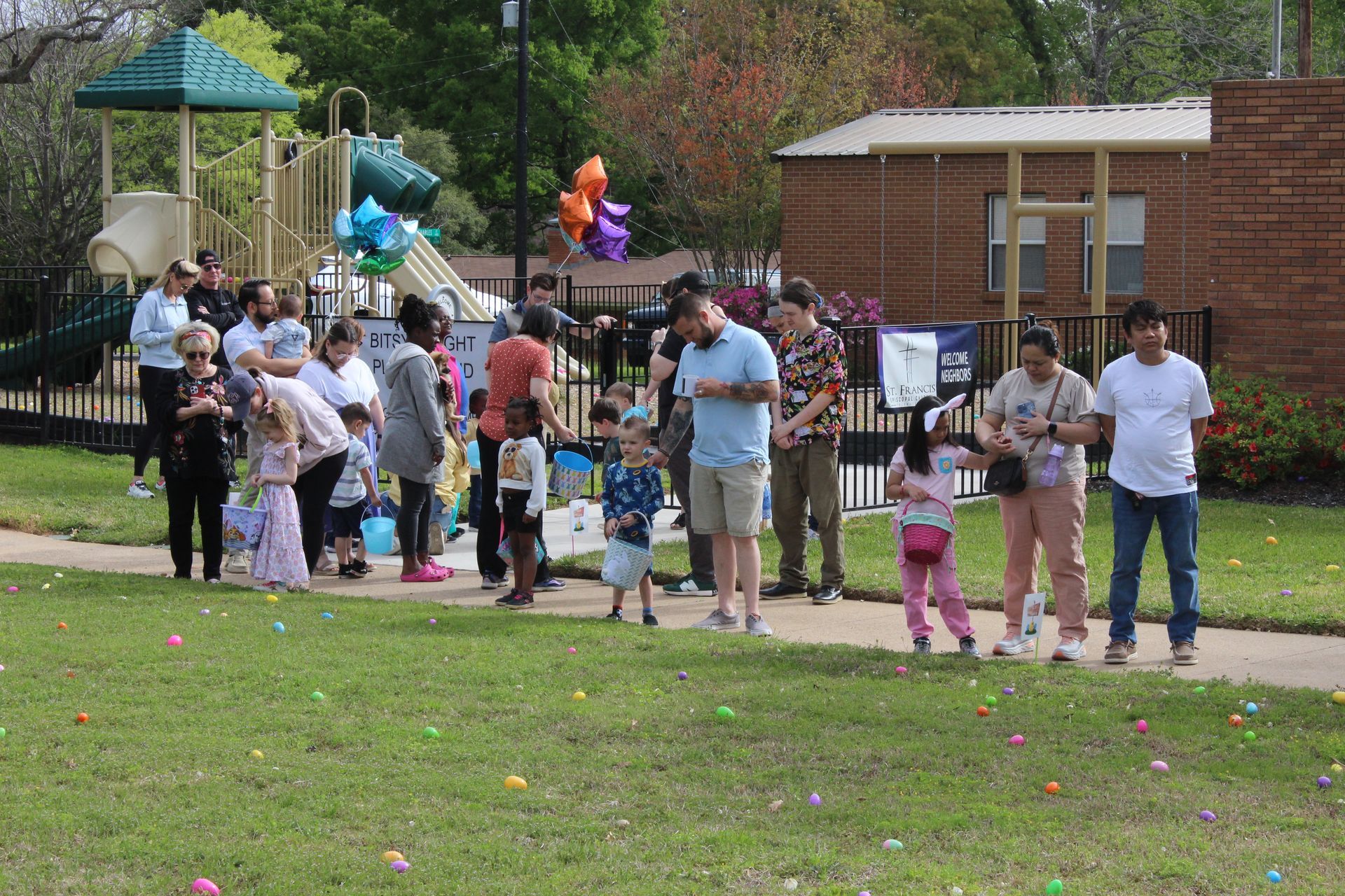 People, including children, gather outside for an Easter egg hunt on a grassy area near a playground and building.