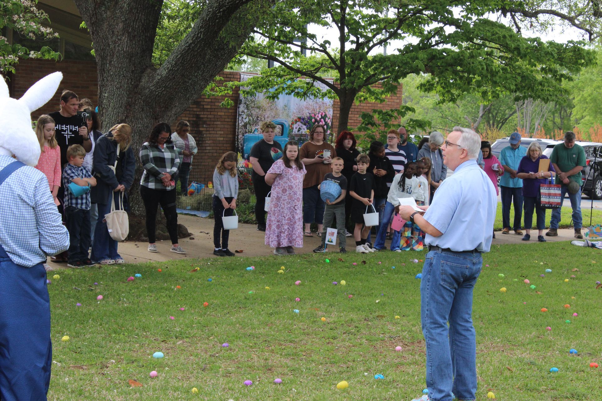 Group of people, including children, gathered outside for an Easter egg hunt, with a person in an Easter bunny costume.