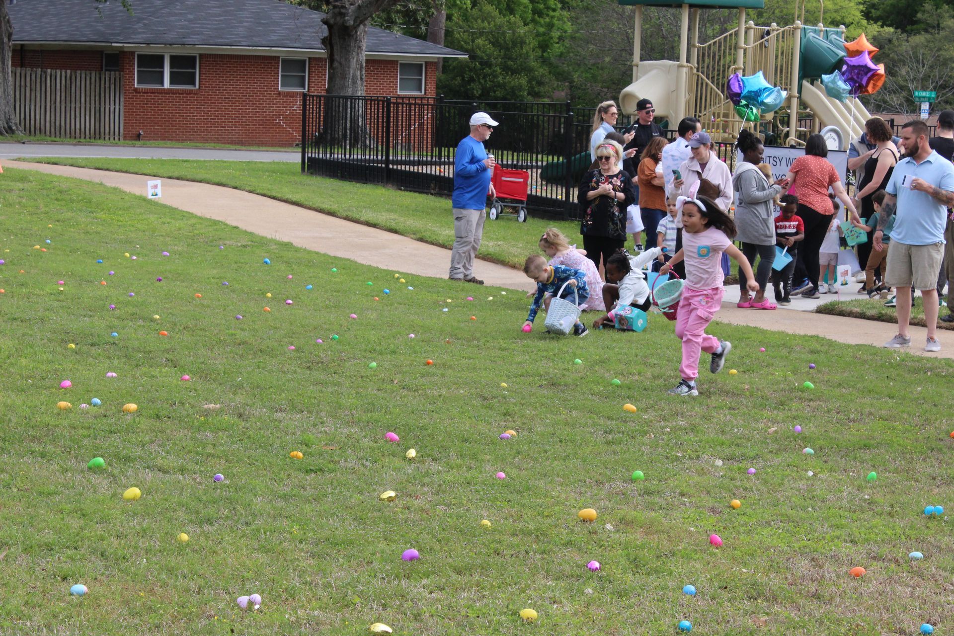 Children hunt for Easter eggs on a grassy field; adults watch near a playground.